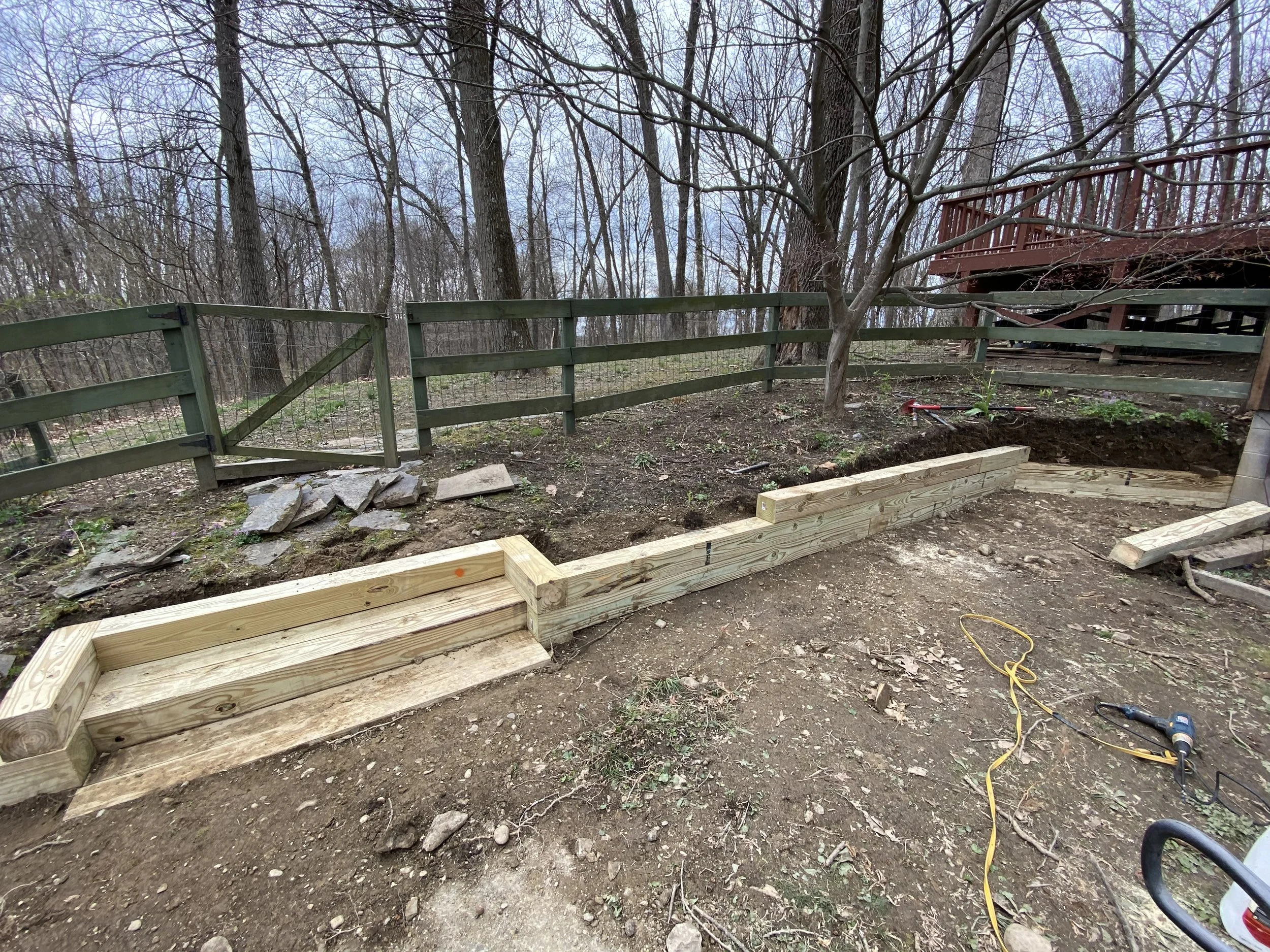 Wooden retaining wall construction in a wooded backyard near a green fence and deck.