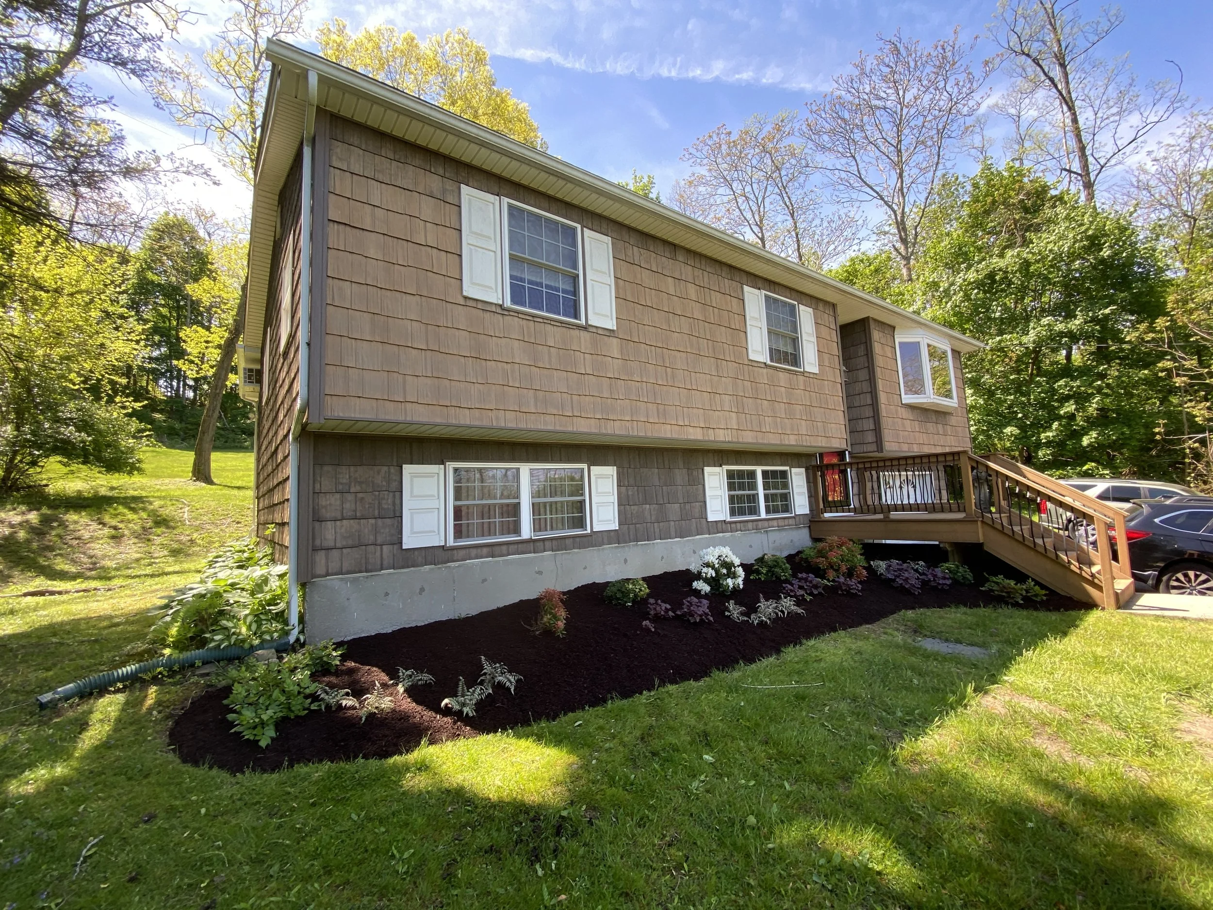 A suburban split-level home with brown wooden siding, white window shutters, and a sloped green lawn. The house features a landscaped garden with flowers near the front entrance and a wooden staircase leading to the front door. There are several trees and parked cars in the background.