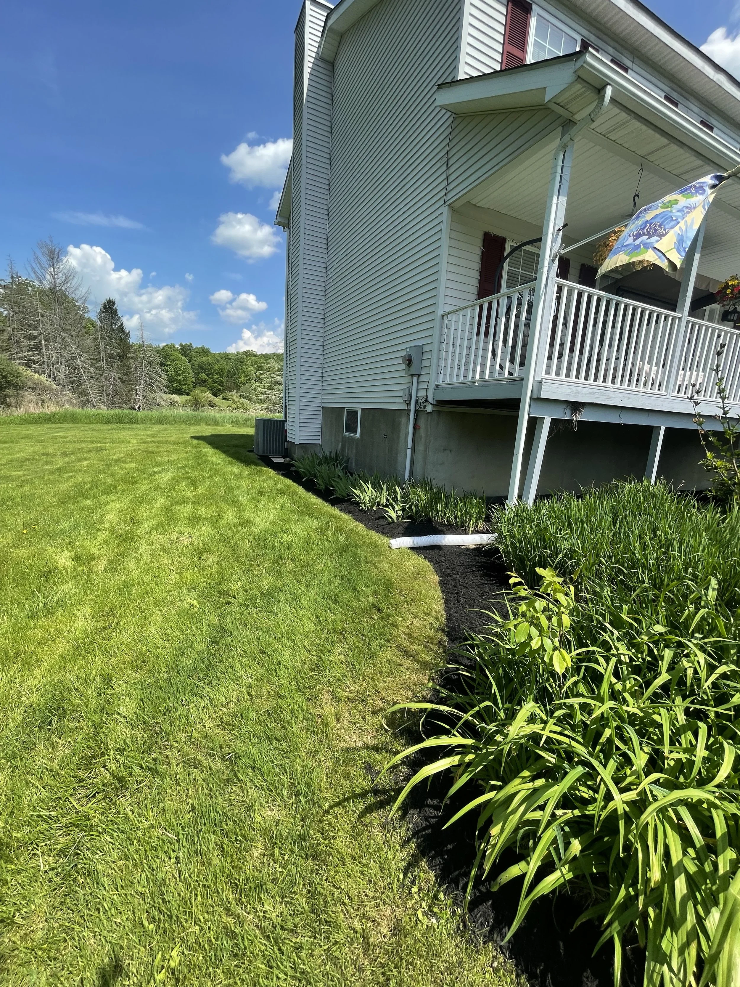 Side view of a white house with a porch, surrounded by green lawn and landscaped garden, under a blue sky with scattered clouds.
