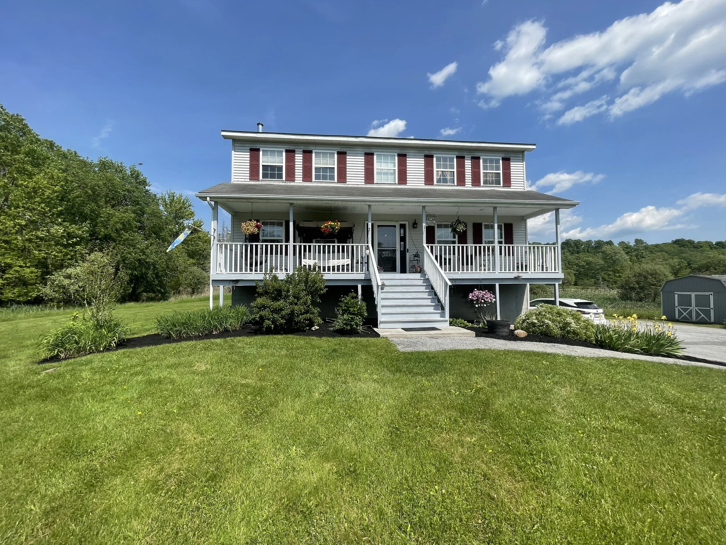 Two-story house with a covered porch, red shutters, and a landscaped front yard featuring flowers and shrubs. A driveway with a shed is visible to the right, under a partly cloudy sky.