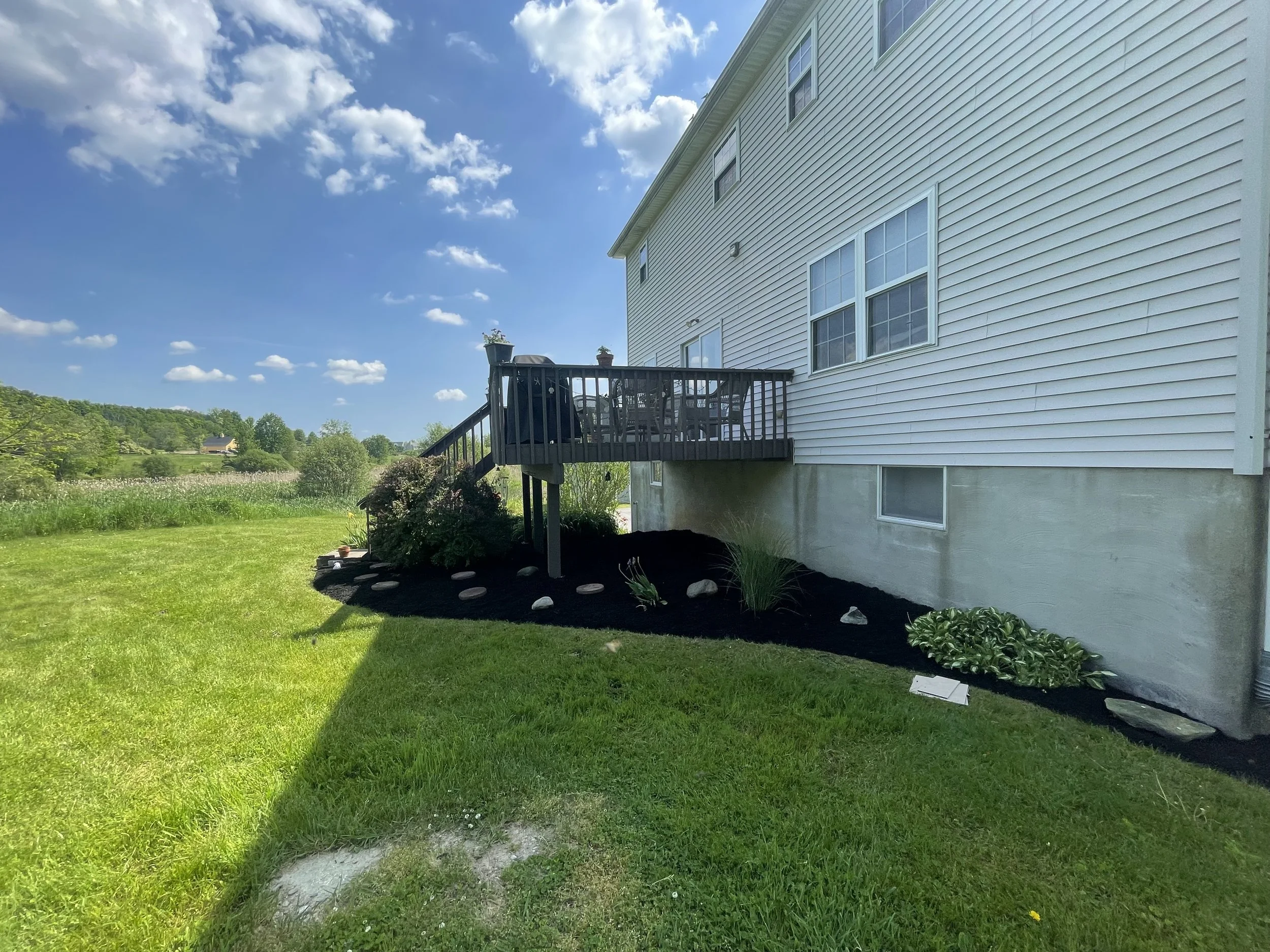 Side view of a house with white siding and a wooden deck, surrounded by a well-maintained garden and green lawn. The sky is clear with a few clouds.