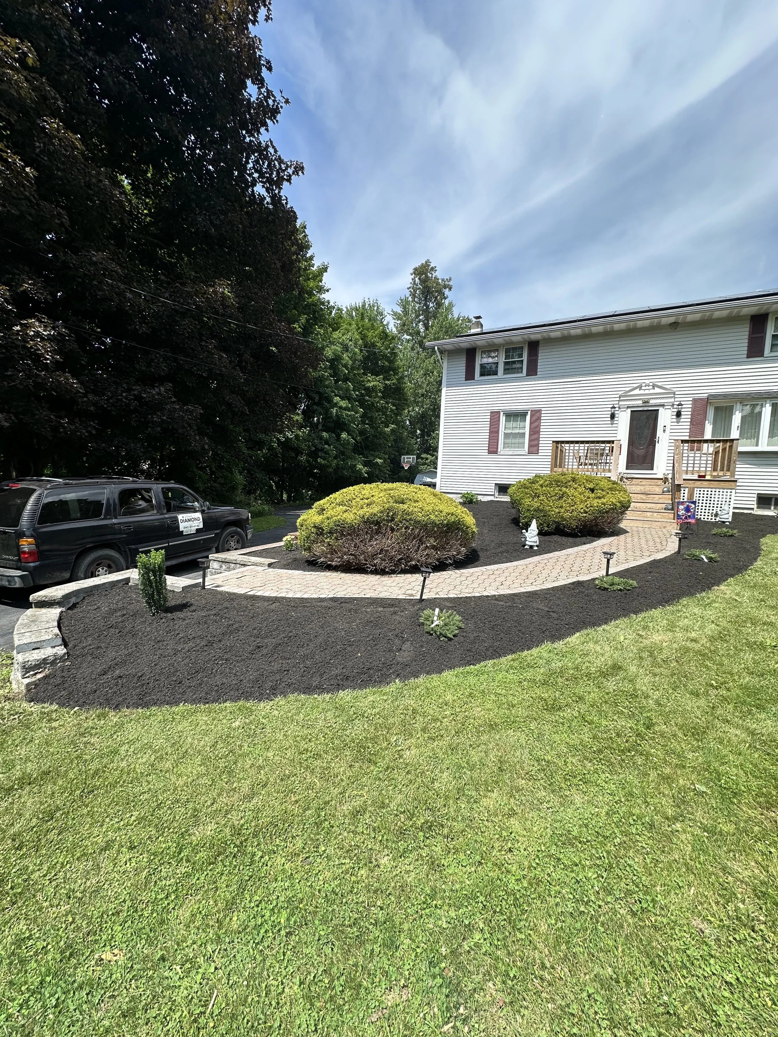 A well-manicured front yard with green grass, and neatly mulched landscaping beds with small plants and bushes. A brick pathway leads to the front door of a white house with burgundy shutters. To the left, there is a parked black SUV. Tall trees and 