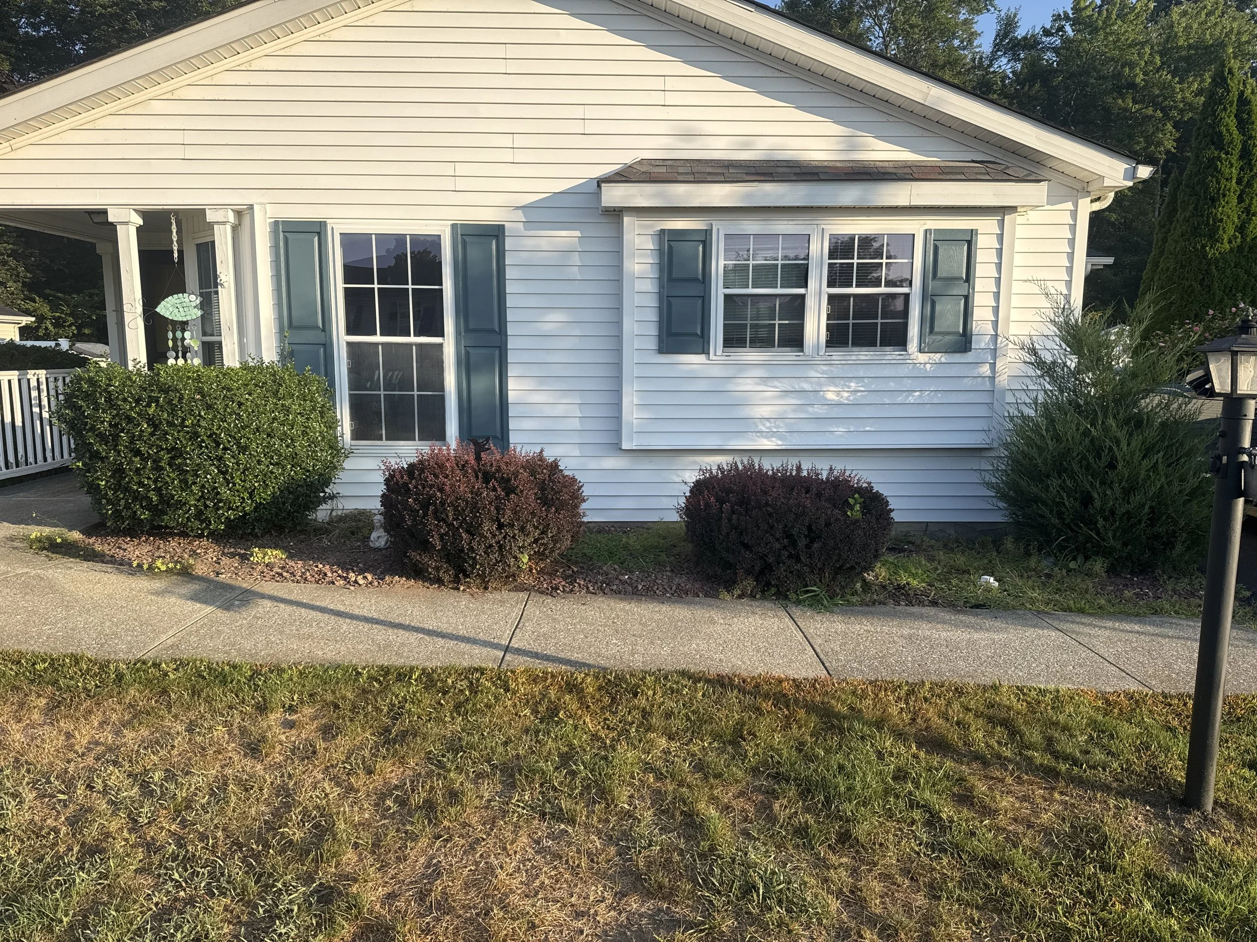 Front view of a white house with blue shutters, a bay window, and a porch with a hanging decoration. There are bushes and landscaping along the sidewalk.