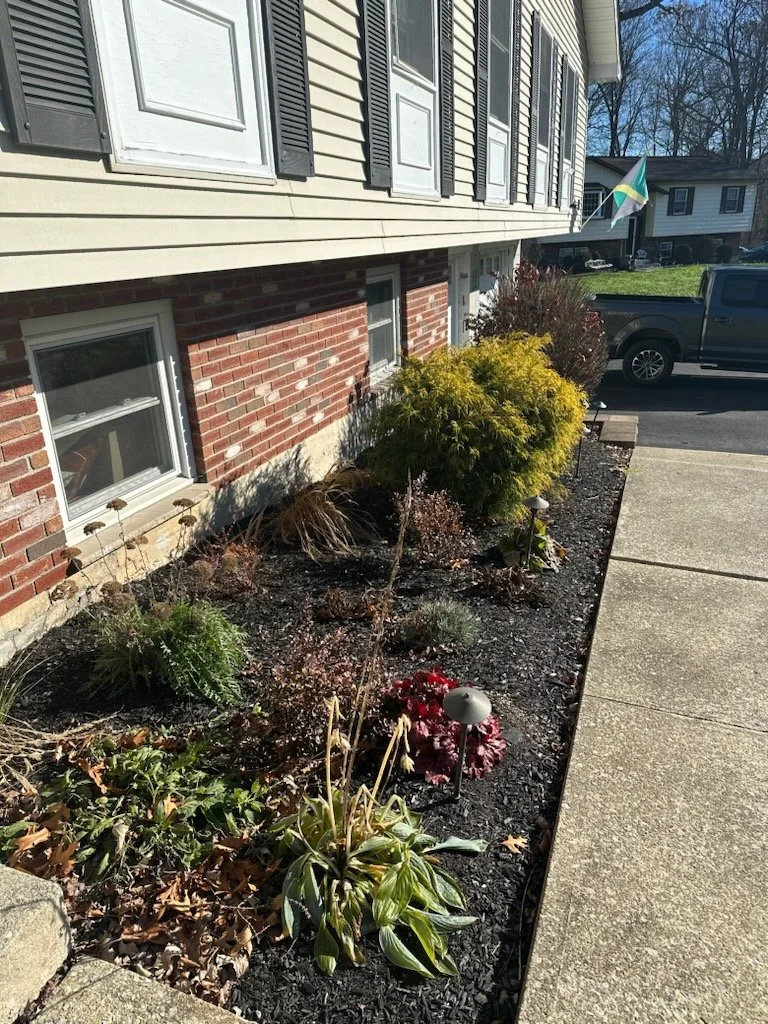 A residential house with beige siding and black shutters, a garden bed with plants and mulch, and a concrete sidewalk in the foreground. A flag is visible in the background.