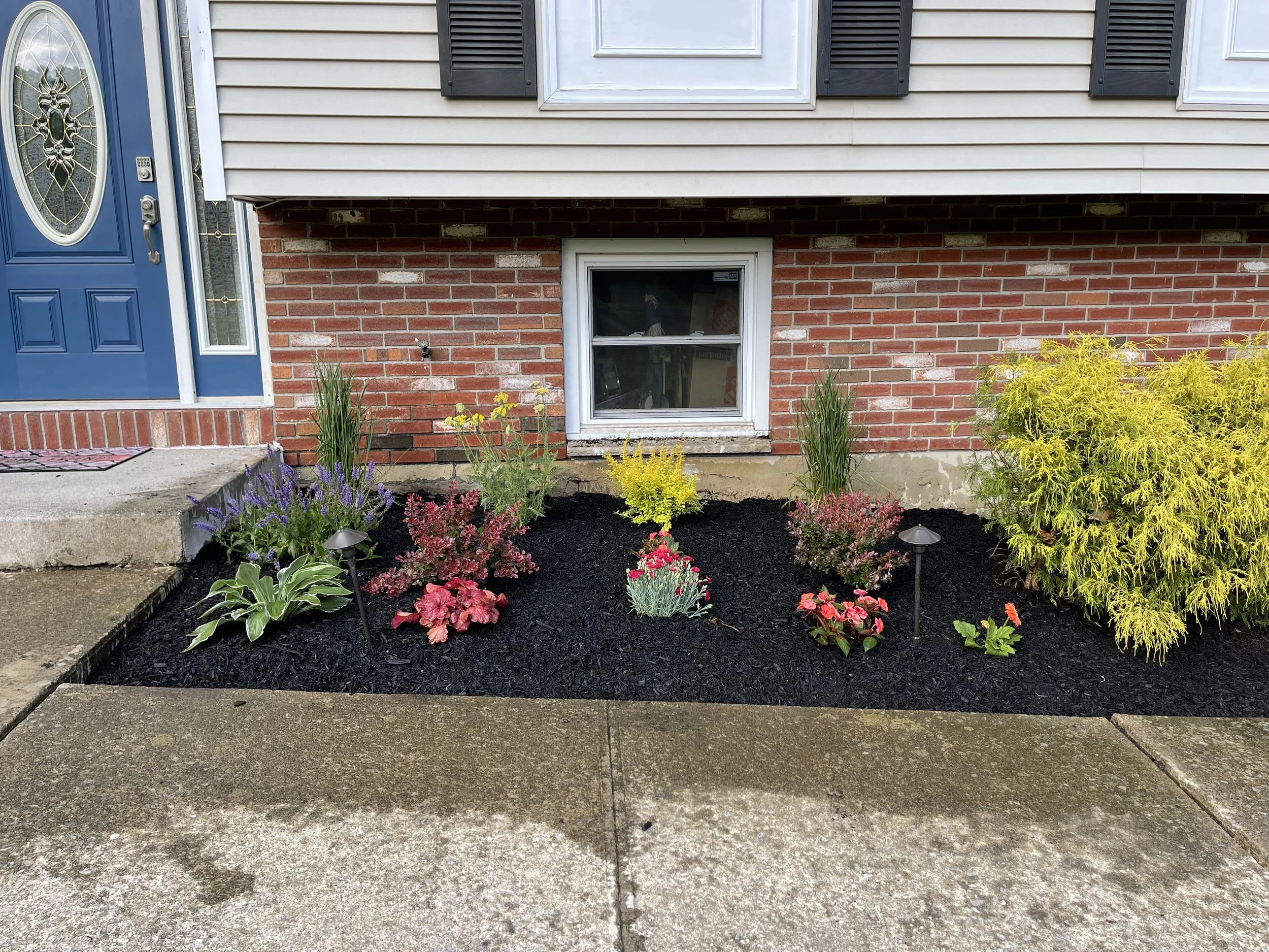 Front yard with flower bed of various colorful plants, small landscape lights, and a concrete sidewalk in front of a brick house with a blue door.