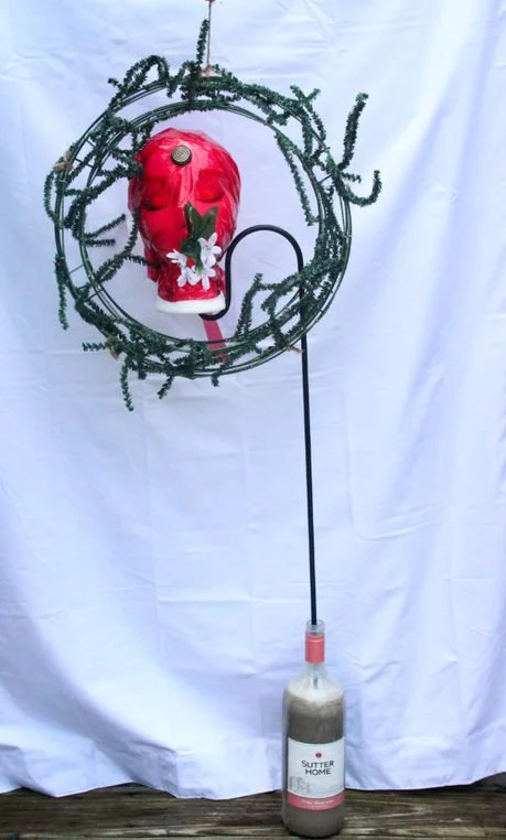 Title: Coriander:

An empty wine bottle, hardened with cement, becomes the body of this piece—heavy, sealed, and immovable. From it rises a foam head wrapped tightly in red cellophane, its surface suffocated in a translucent veil. A wreath encircles 