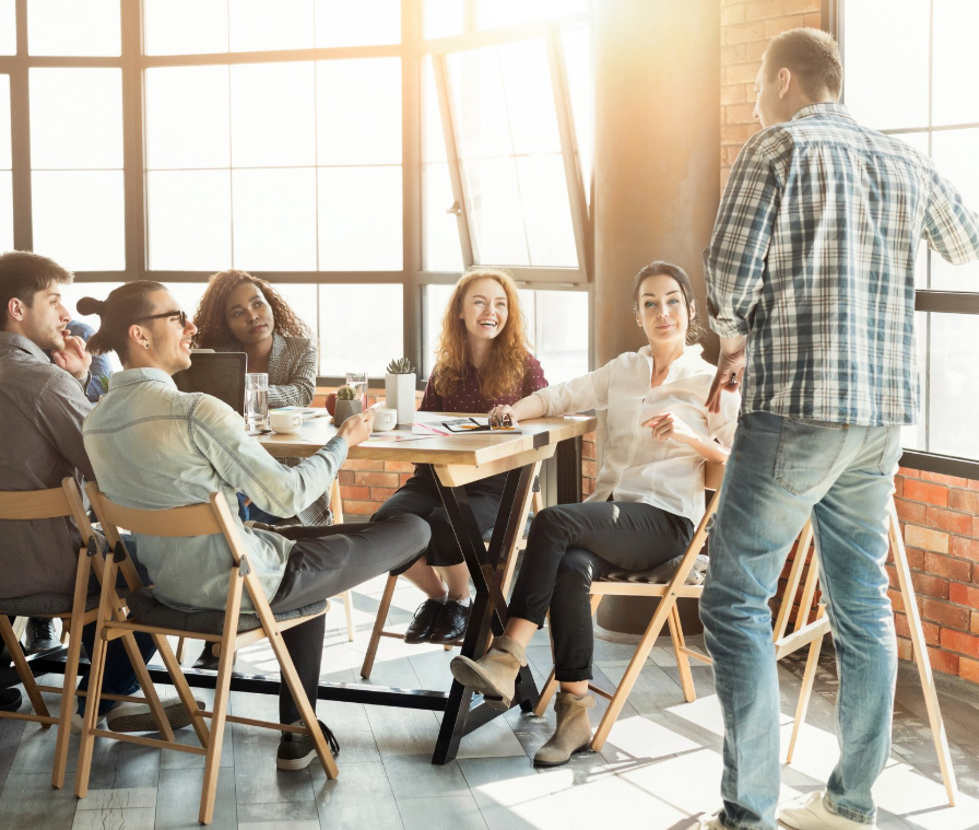 A group of six diverse young adults in a business meeting or workshop in a bright, modern office space with large windows, listening to a man standing and speaking.