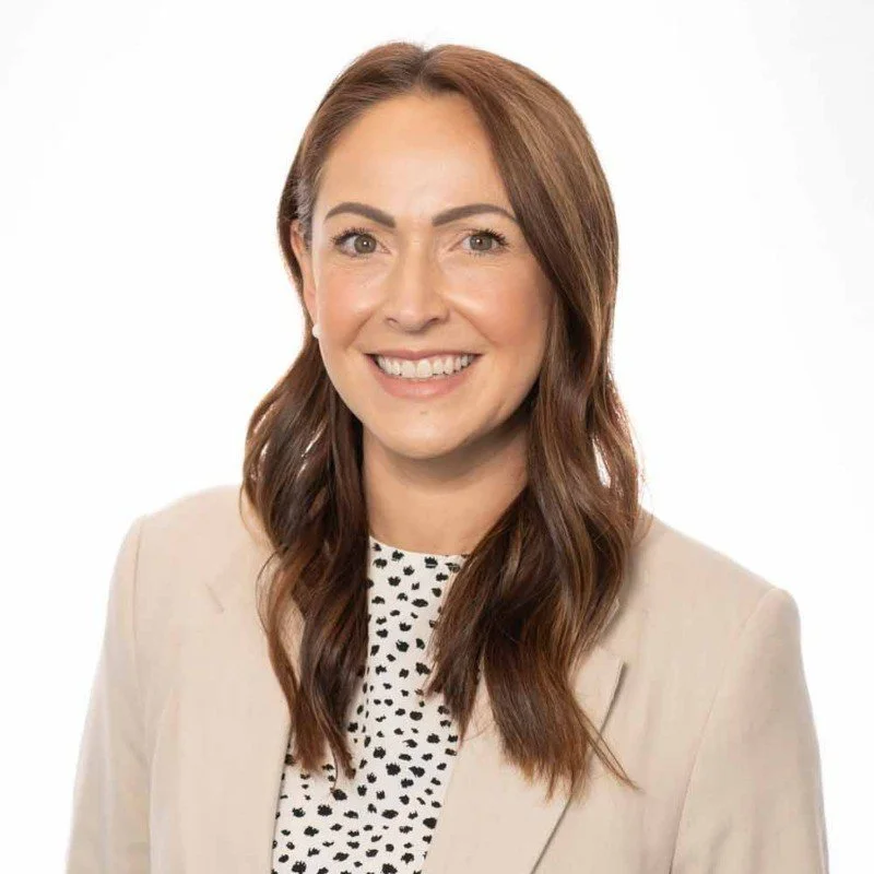 A woman with long brown hair smiling, wearing a beige blazer over a white top with black polka dots, against a white background.