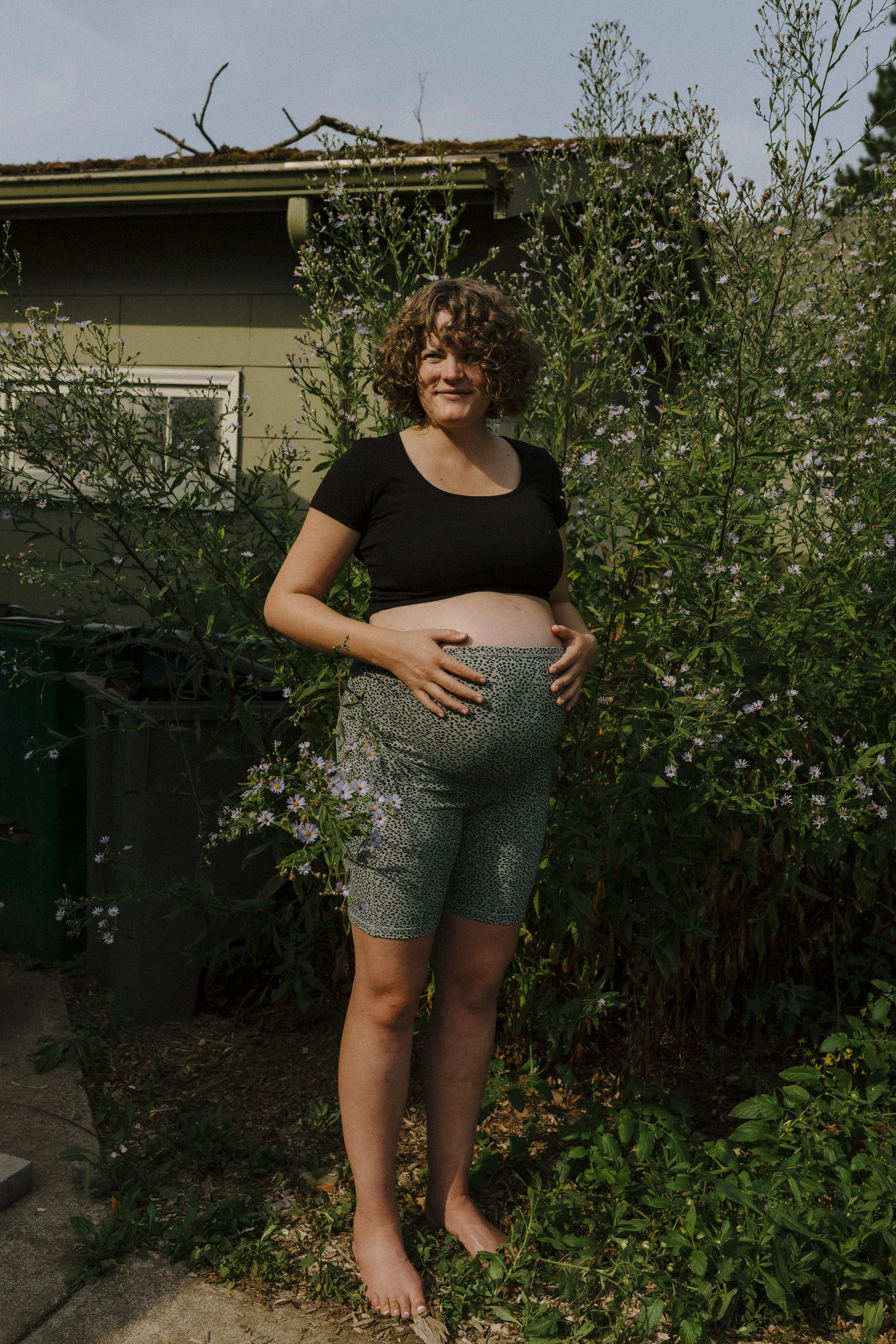 A woman with curly hair is standing outdoors near bushes with purple flowers, gently holding her pregnant belly and smiling, with a house in the background.