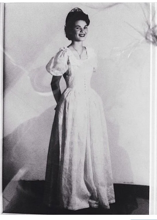 A woman dressed in vintage attire with a long, flowing dress and puffed sleeves, standing and smiling for the camera.
