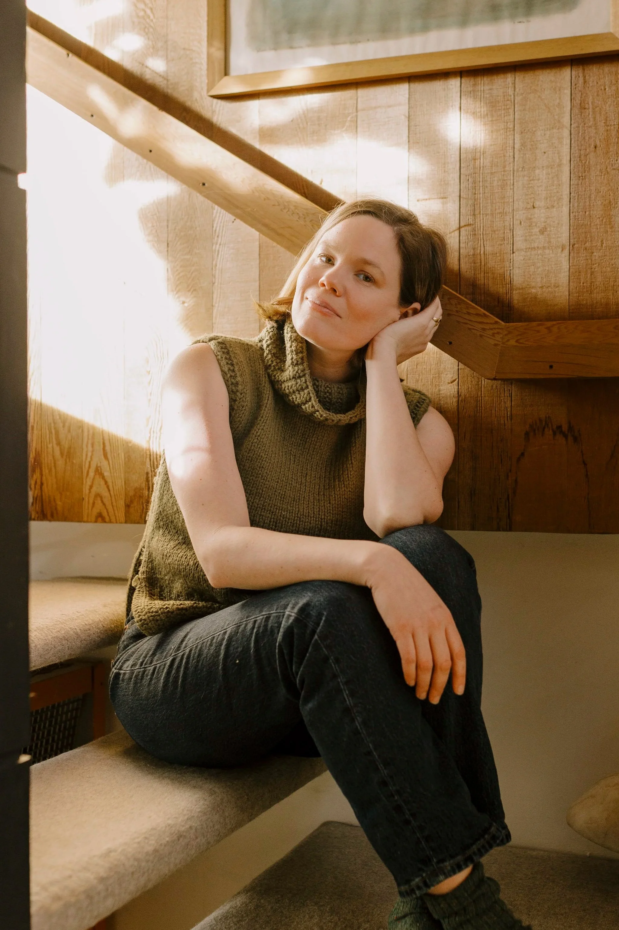 A woman with short brown hair sitting on a beige cushioned seat against a wooden wall, resting her head on her hand, wearing a brown sleeveless sweater and dark pants.
