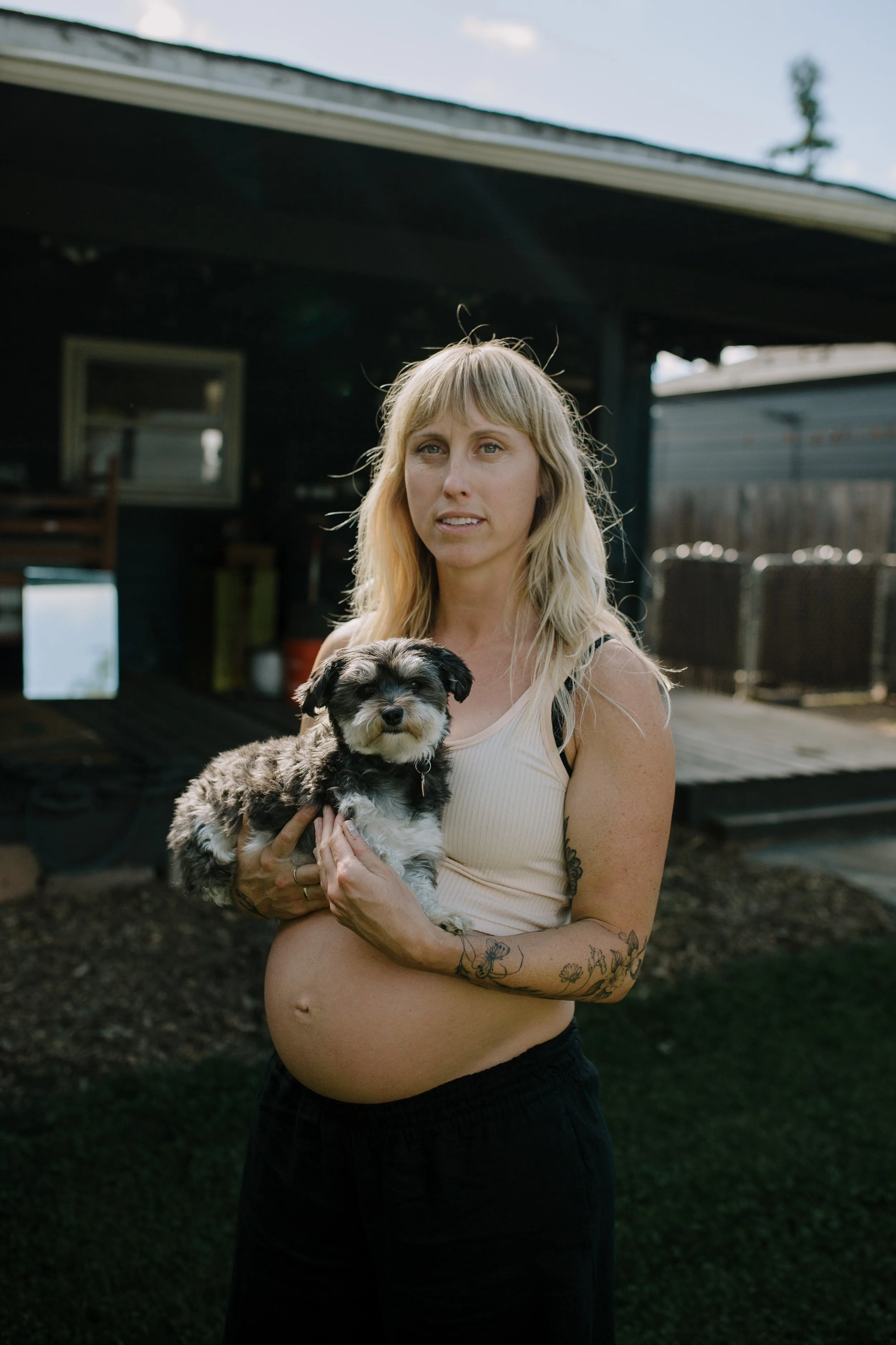 A woman with long blonde hair, tattoos on her arms, and wearing a beige top is holding a small black and gray puppy. They are standing outside in front of a dark building with a yard, some outdoor furniture, and a blue sky in the background.