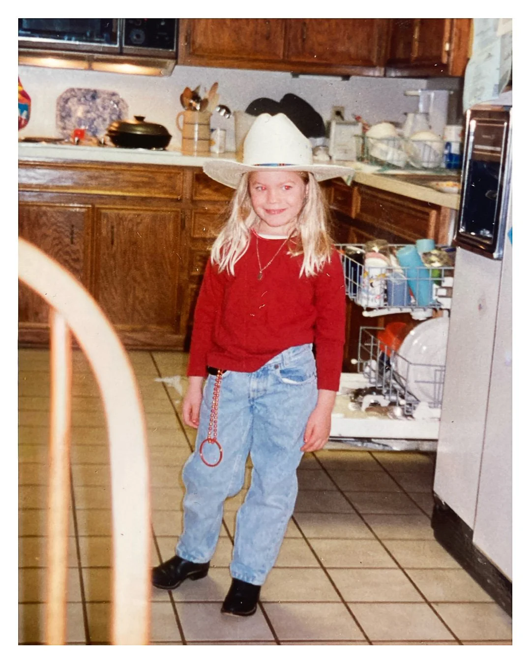 A young girl wearing a cowboy hat, red sweater, light blue jeans, and black boots, standing in a kitchen.
