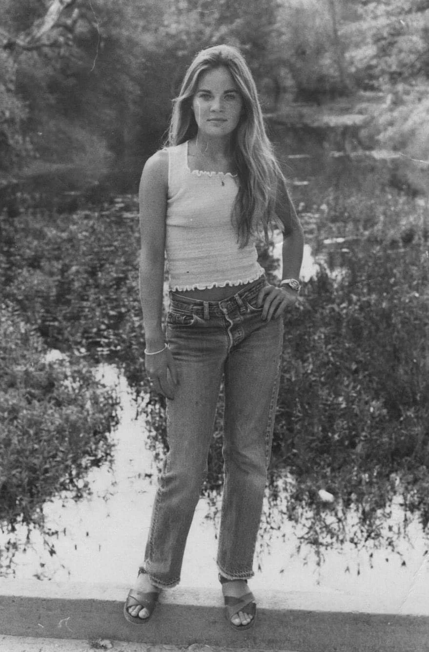 A young woman with long hair stands outdoors near a body of water, wearing a sleeveless top, jeans, and sandals.