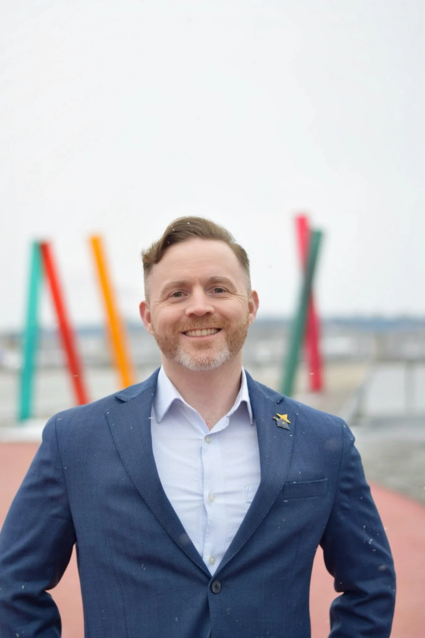 Smiling man in a blue blazer and white shirt standing outdoors with colorful abstract sculptures in the background.