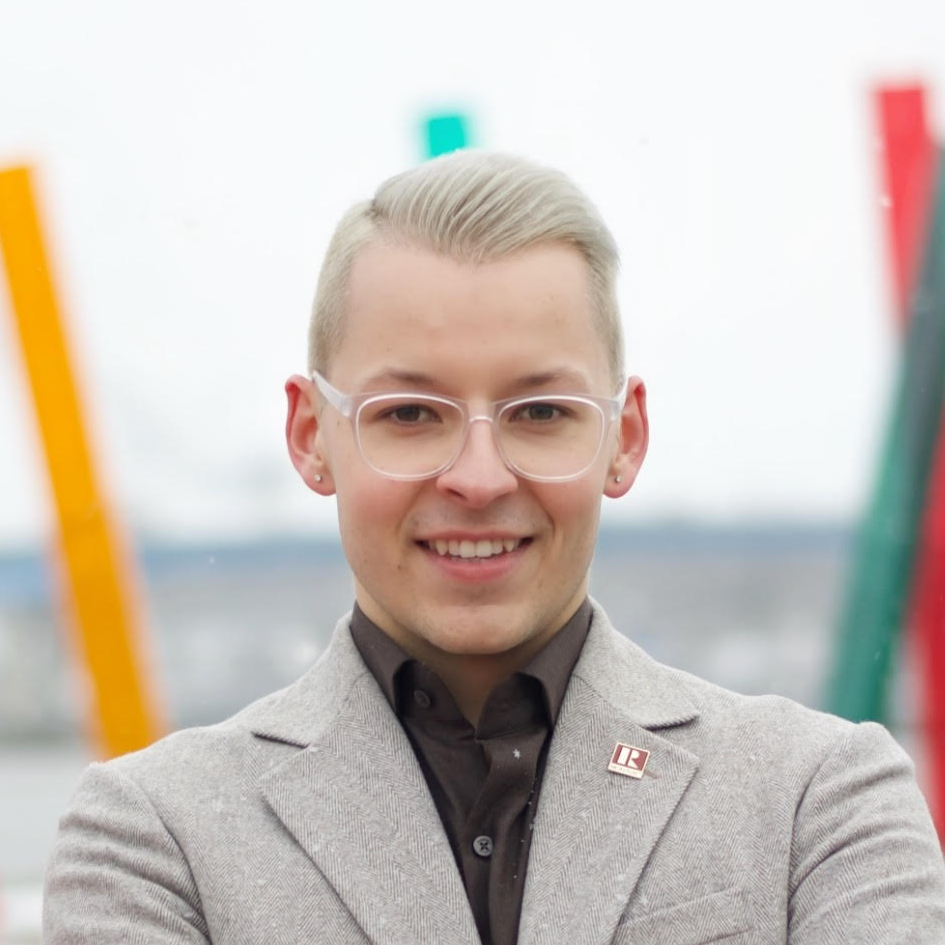 A young man with short blonde hair, clear glasses, and earrings, smiling and wearing a gray blazer over a dark shirt, standing outdoors with colorful abstract structures in the background.