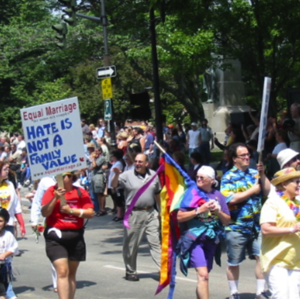 People participating in a parade or protest holding signs supporting equal marriage rights and LGBTQ+ pride, with trees and a crowd in the background.