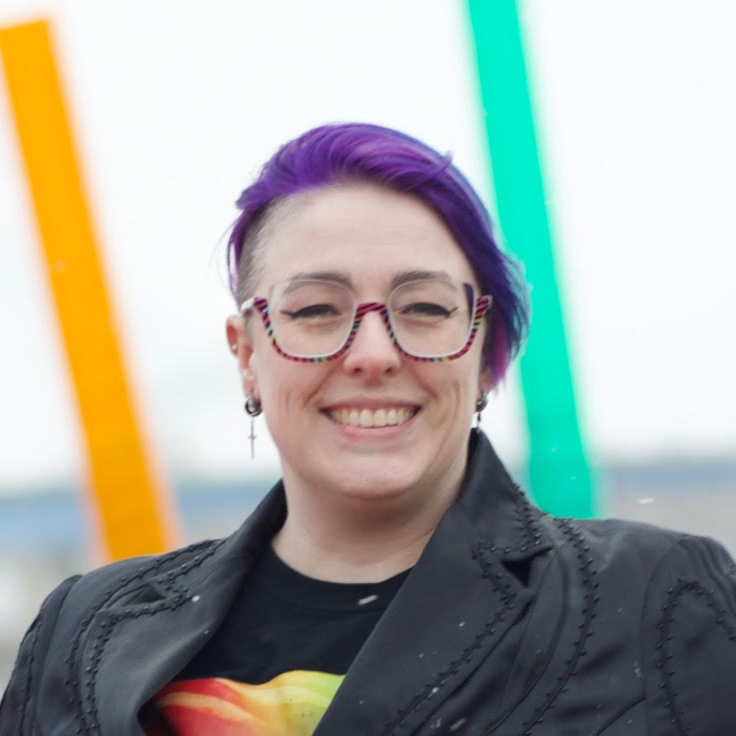 A smiling person with purple hair, wearing glasses with colorful frames, a black jacket, and a graphic T-shirt, standing outdoors with blurred colorful structures in the background.