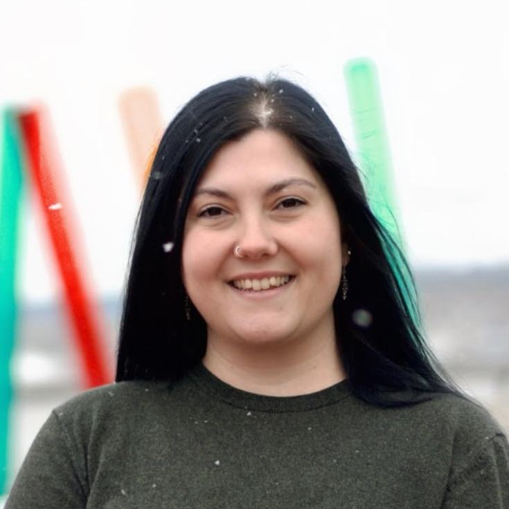 A young person with black hair, smiling, wearing a dark green shirt, standing outdoors with colorful abstract sculptures in the background.