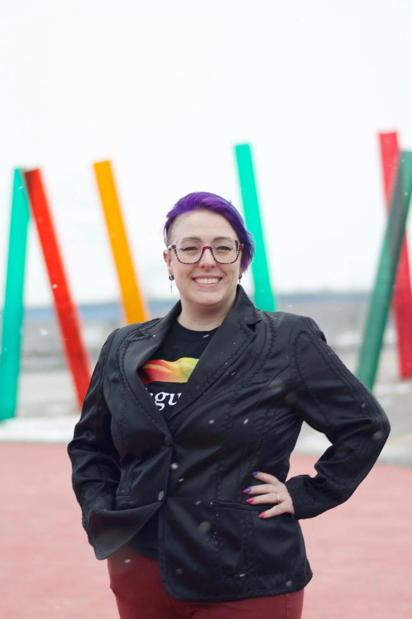 A person with purple hair, glasses, and a black jacket smiling with their hand on their hip outdoors in front of colorful vertical art installation.