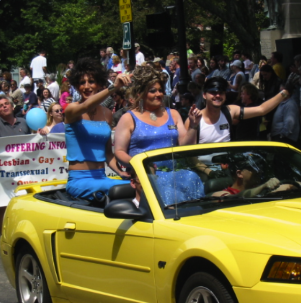 Three people sitting in a yellow convertible car during a pride parade, with a crowd of spectators and a banner in the background.