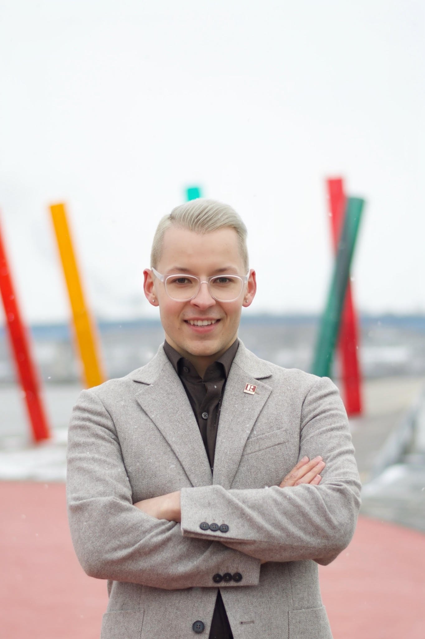 A smiling young man with glasses and blonde hair standing outdoors with arms crossed. He is wearing a grey blazer over a black shirt. There are colorful abstract outdoor sculptures and a body of water in the background.