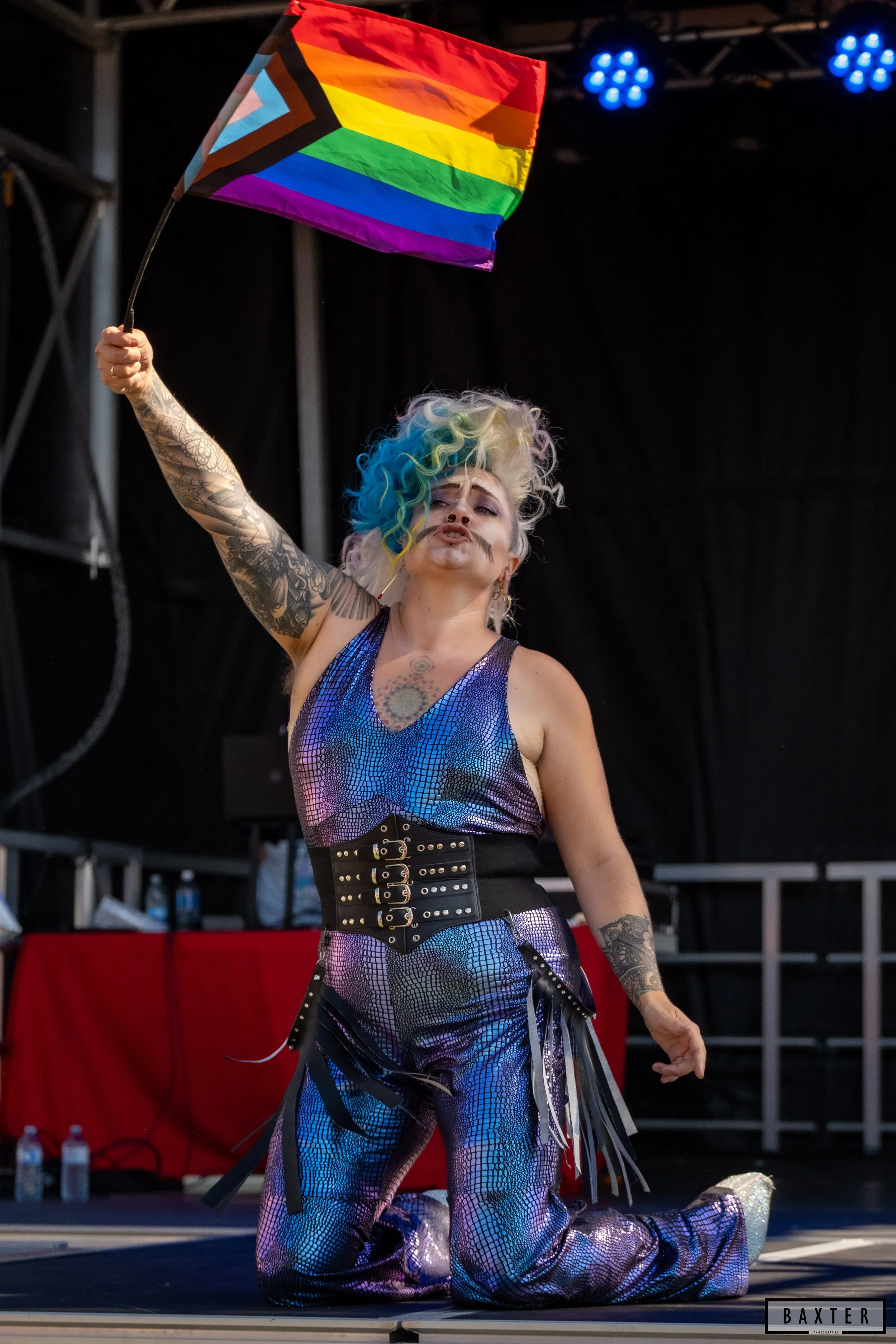 An individual with colourful rainbow hair and tattoos holding a rainbow pride flag on a stage, kneeling with one knee down and lips puckered.