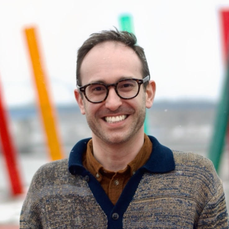 A smiling man with glasses and a beard standing outdoors with colorful vertical poles in the background.