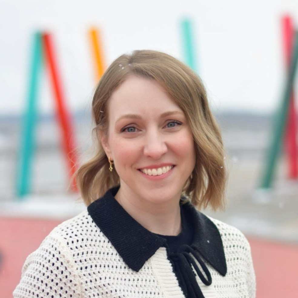 A smiling woman with short, wavy, light brown hair stands outdoors in front of colorful abstract sculptures.