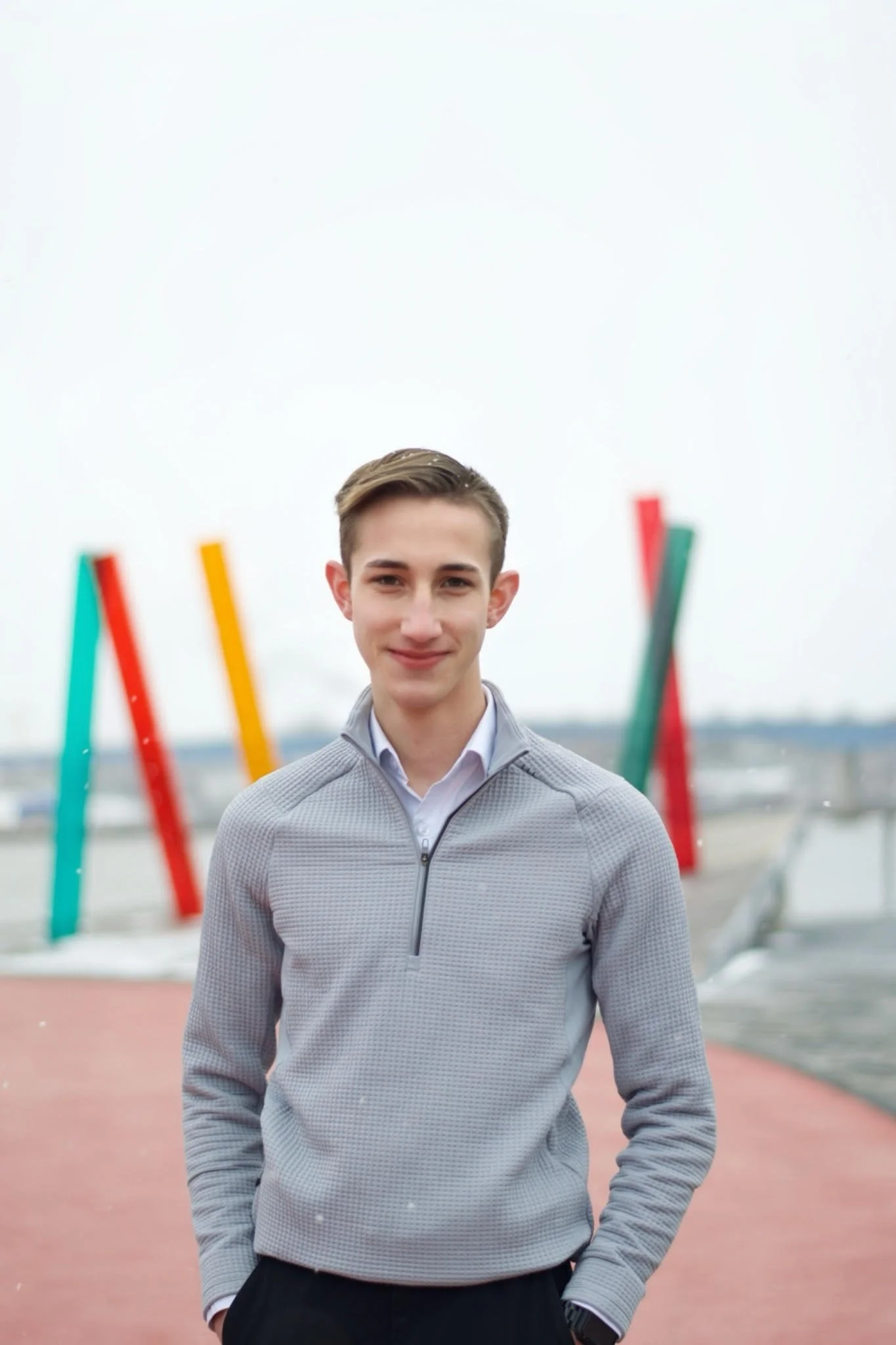 A young man with short brown hair smiling at the camera, standing outdoors with colorful abstract sculptures and an overcast sky in the background.