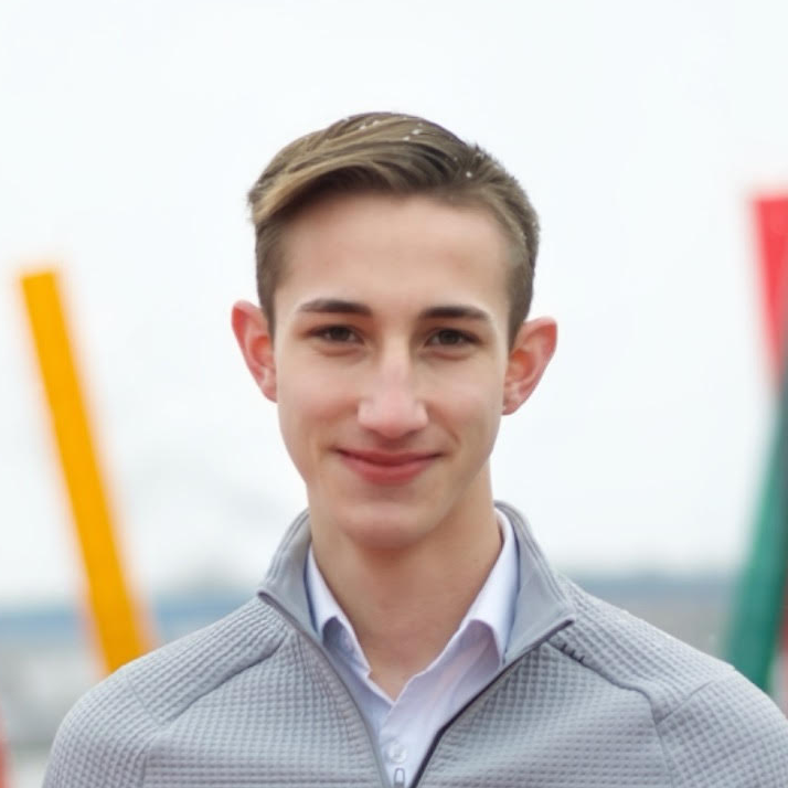 Young man with short, light brown hair and fair skin, wearing a light gray zip-up jacket over a white collared shirt, smiling outdoors with blurred flags in the background.