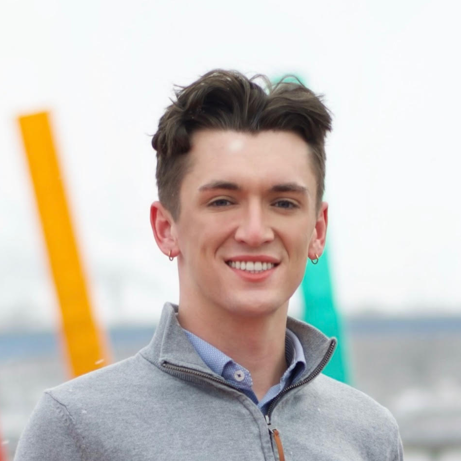 Young man with short brown hair, earrings, wearing a gray zip-up sweater, smiling outdoors near water with colorful vertical structures in the background.