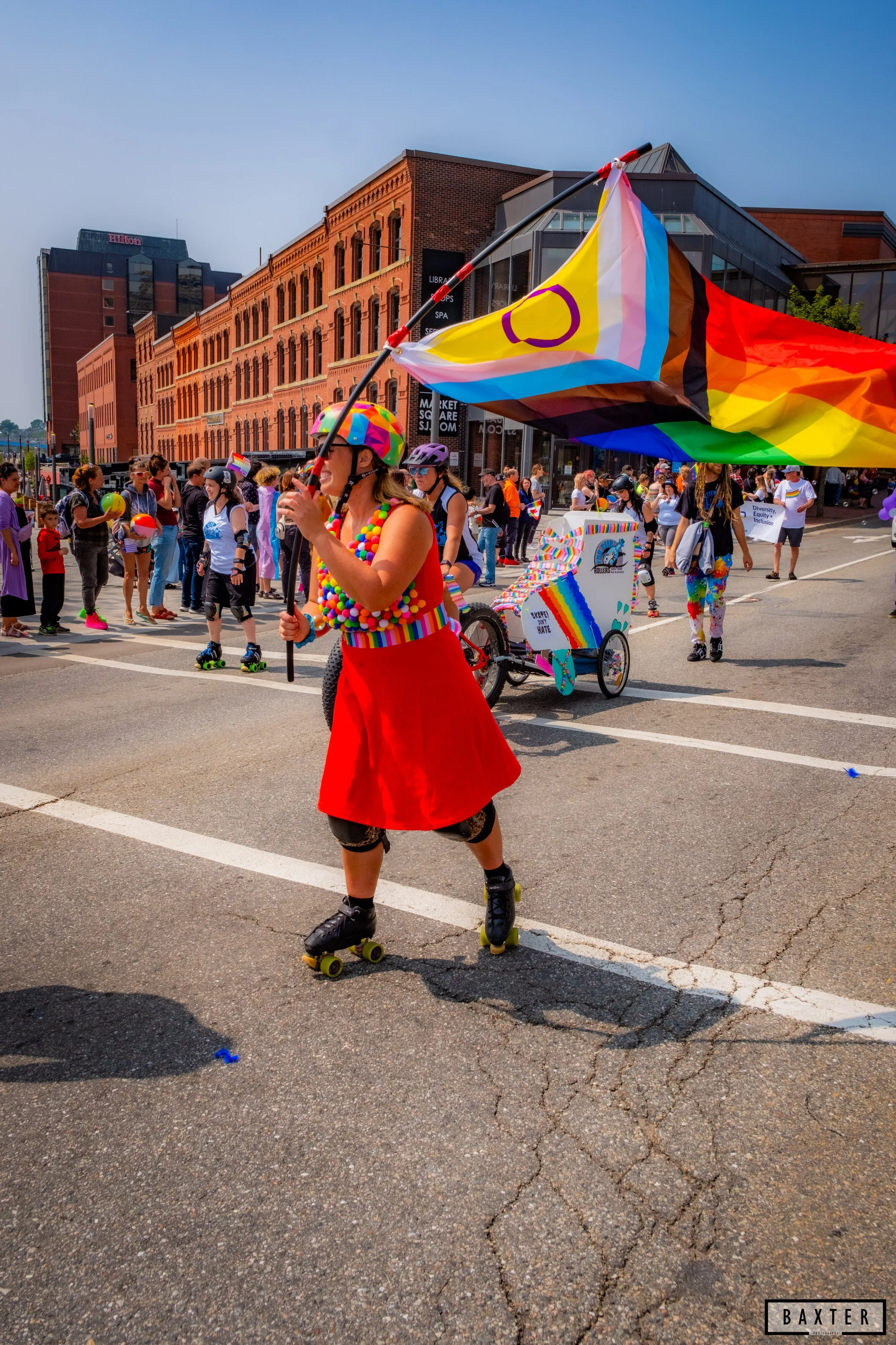 Person on roller skates wearing a rainbow-colored helmet and red dress, holding a rainbow pride flag, participating in a pride parade on a city street with many spectators and rainbow decorations.