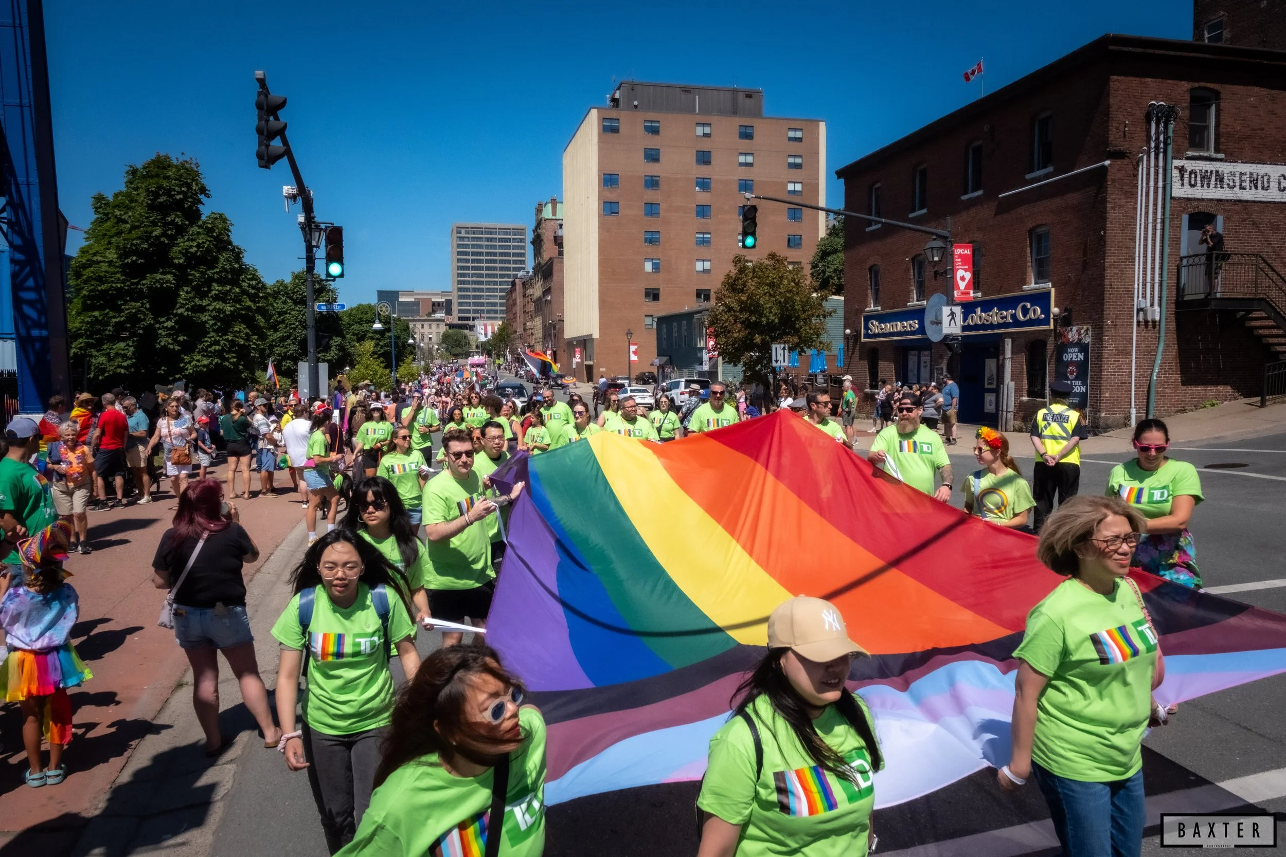 Group of people participating in a pride parade, holding a large rainbow flag on a city street.