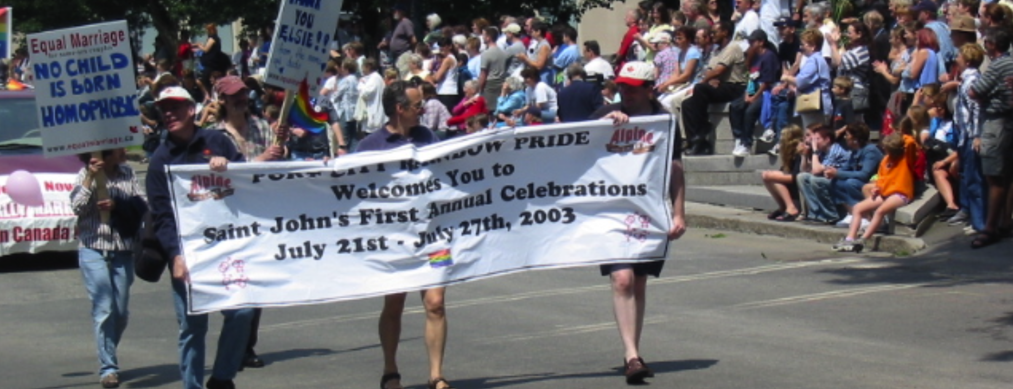 Crowd participating in a Pride parade, with some holding signs advocating for LGBTQ+ rights. The main banner welcomes attendees to Saint John's First Annual Celebrations, July 21-27, 2003.