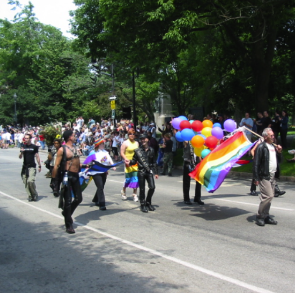 People participating in a pride parade, carrying rainbow flags and balloons, with trees and spectators in the background.