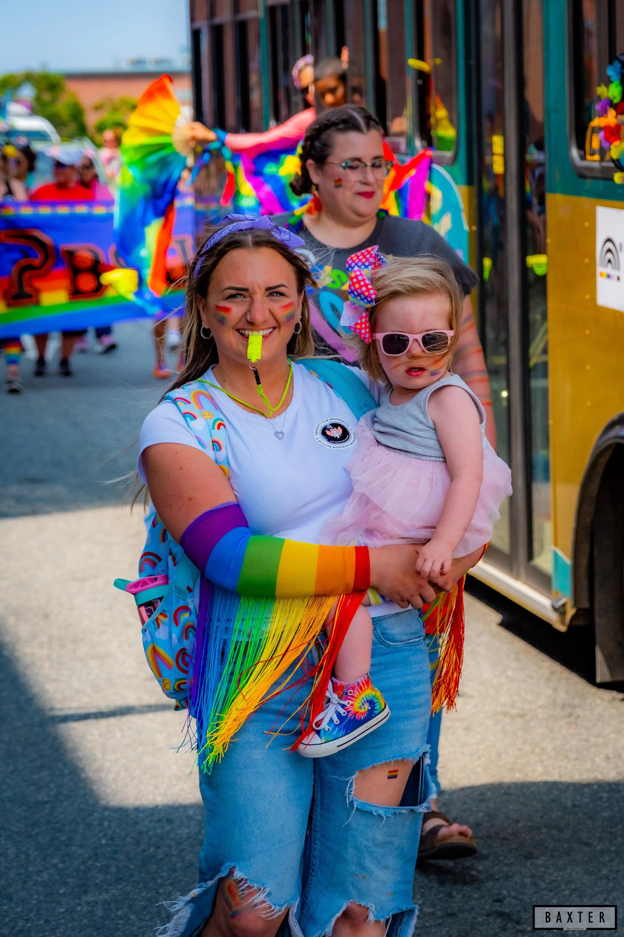 People celebrating Pride parade, including a woman with rainbow-colored accessories and a young girl with rainbow tie-dye shoes and rainbow makeup.