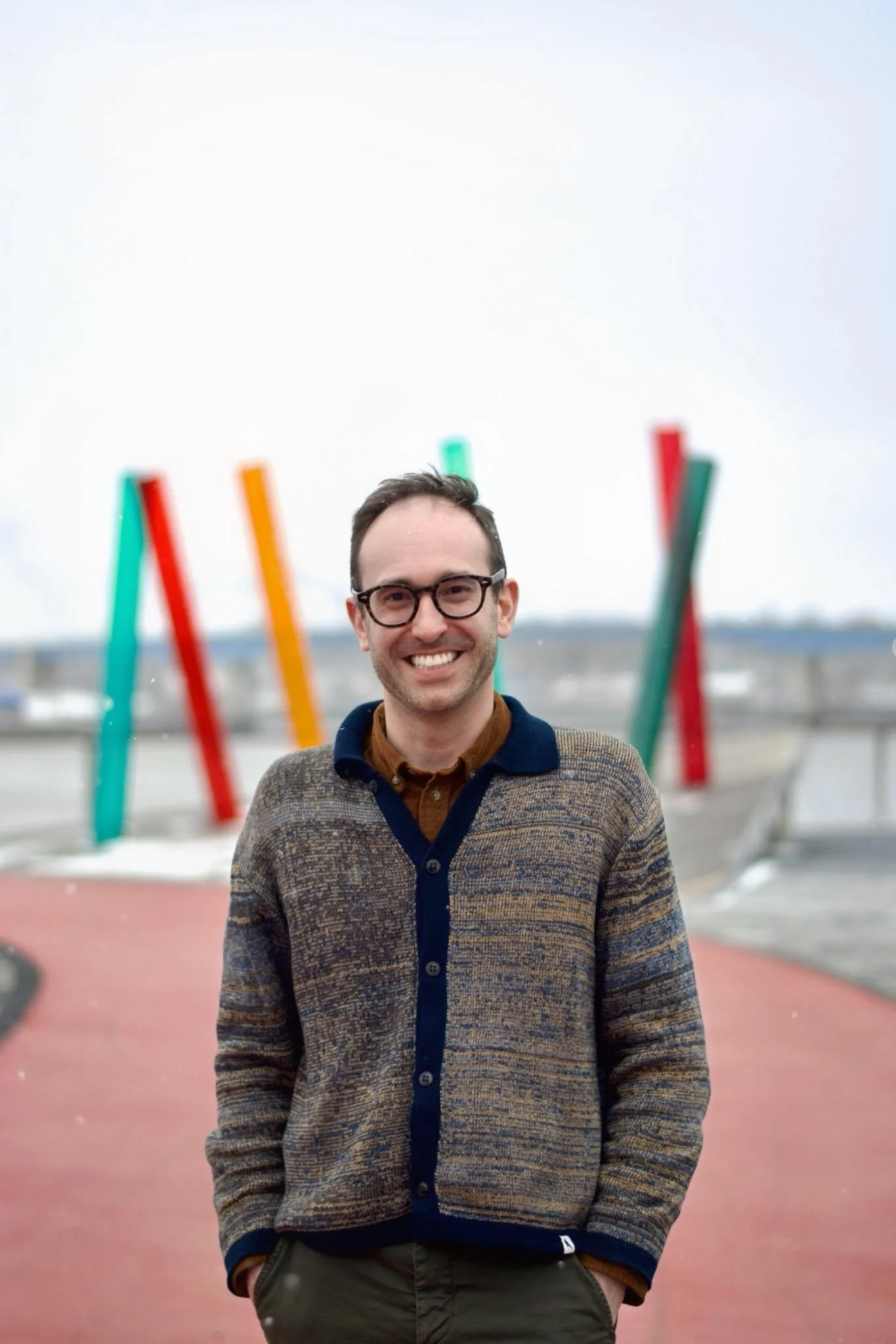 A man with glasses and a patterned sweater smiling outdoors in front of colorful vertical art sculptures and a cloudy sky.