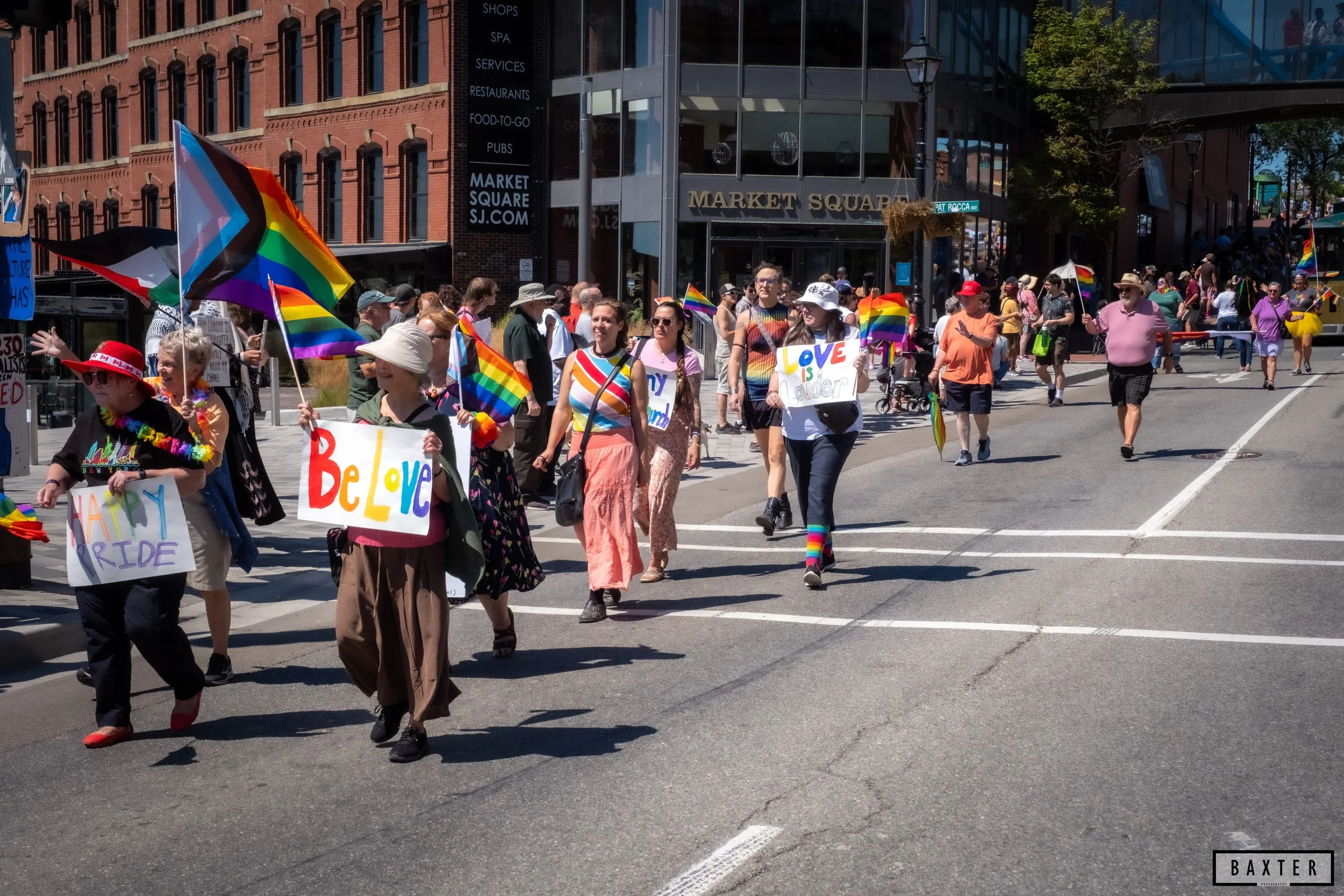 People participating in a pride parade, holding rainbow flags and signs with messages of love and equality, walking in the street outside a building labeled Market Square, with some individuals wearing rainbow-themed clothing and accessories.