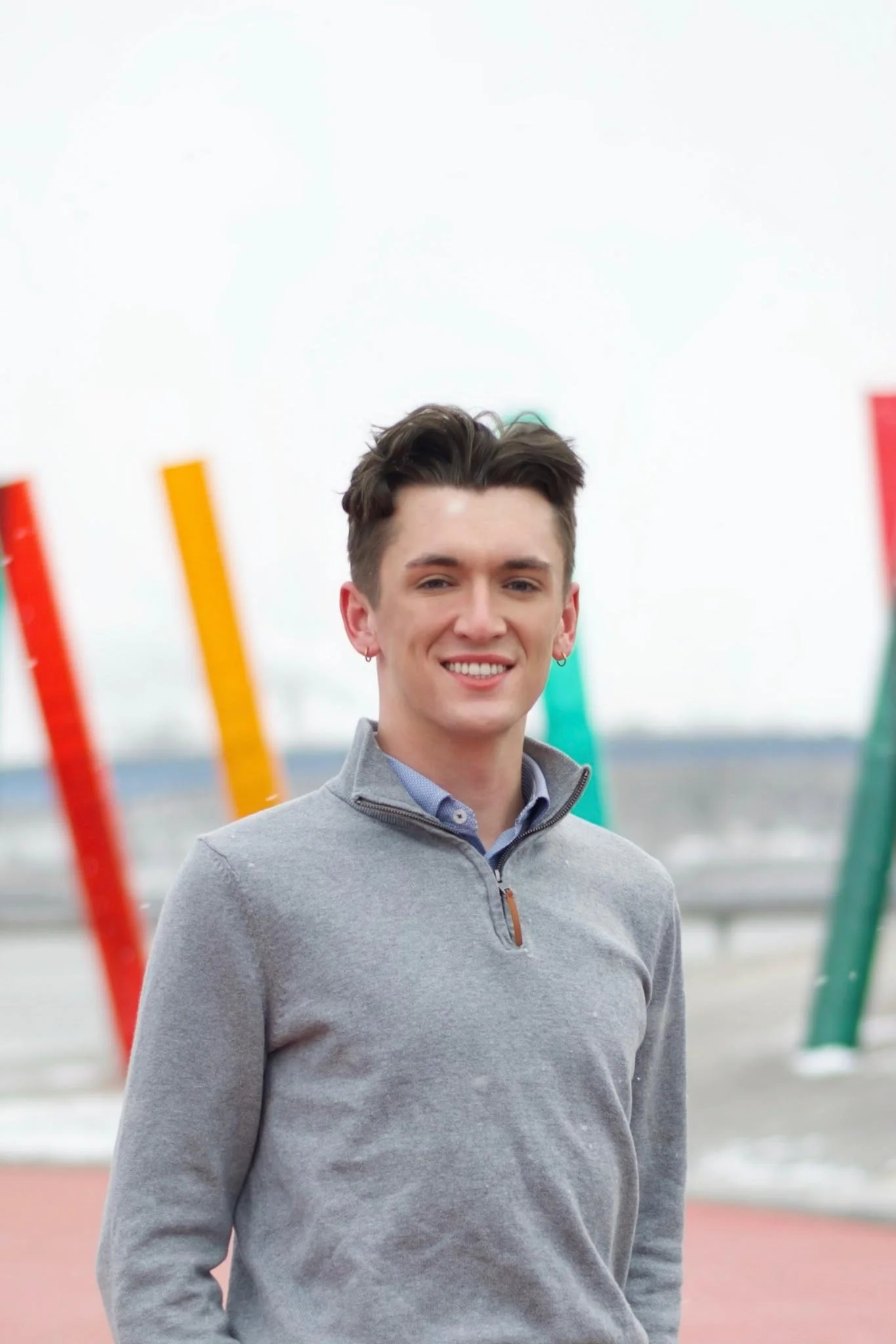 Young man with dark hair styled back, wearing a gray quarter-zip sweater over a blue collared shirt, standing outdoors near colorful abstract structures on a cloudy day.