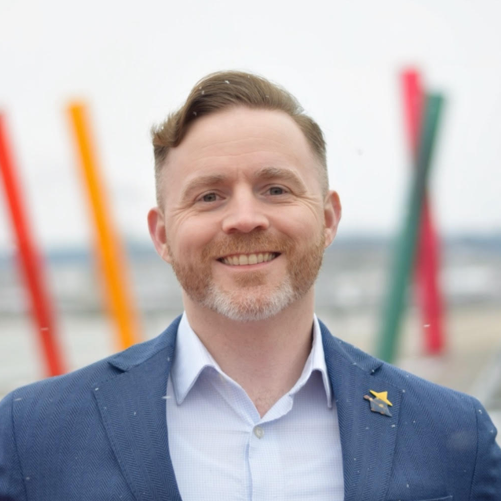 A smiling man with a beard wearing a blue suit jacket and white shirt, standing outdoors with colorful abstract sculptures in the background.