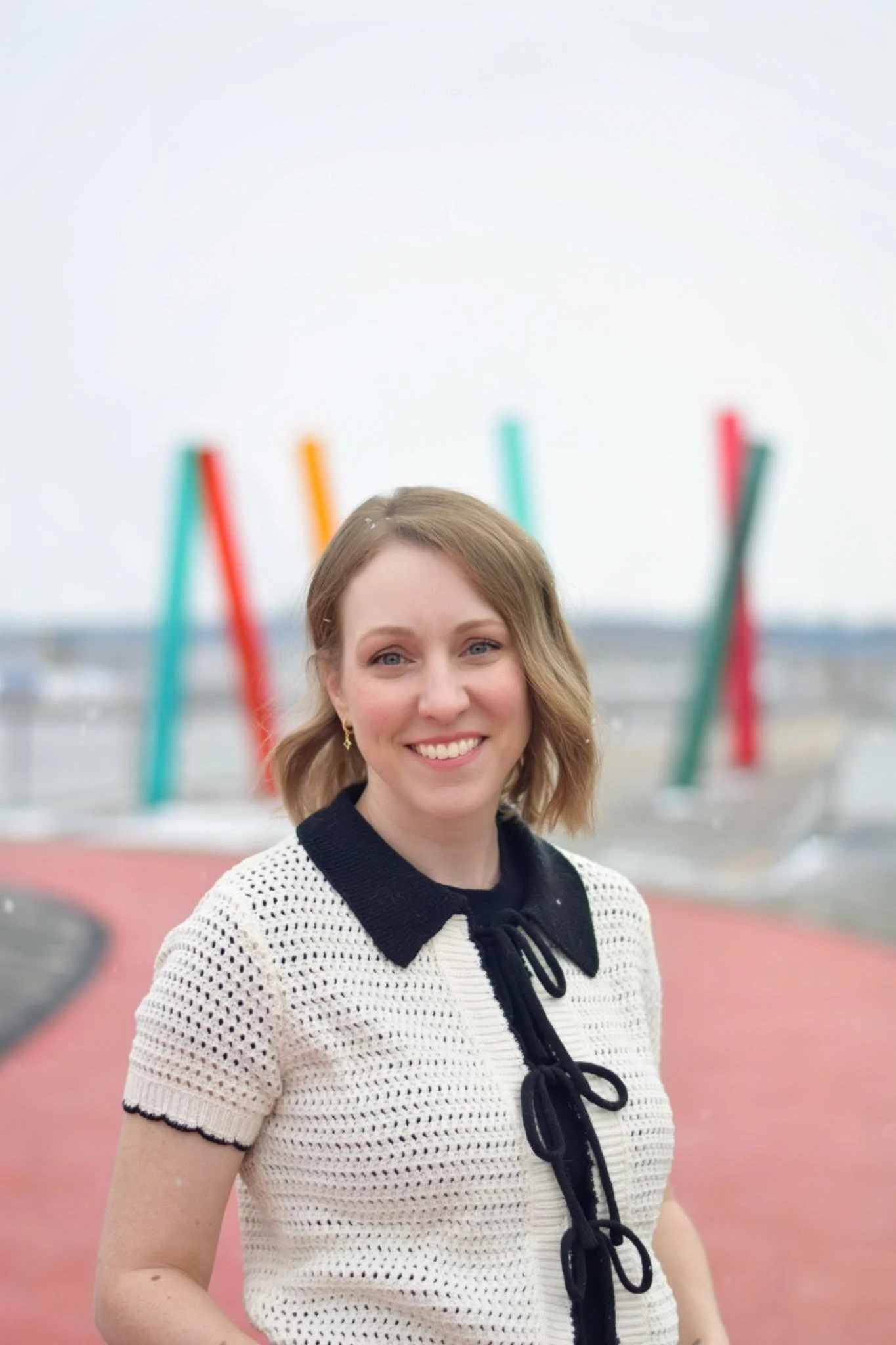 A smiling woman with shoulder-length blonde hair, wearing a cream-colored crocheted top with black accents on the collar and bows, stands outdoors on a pink pathway with colorful abstract sculptures in the background.