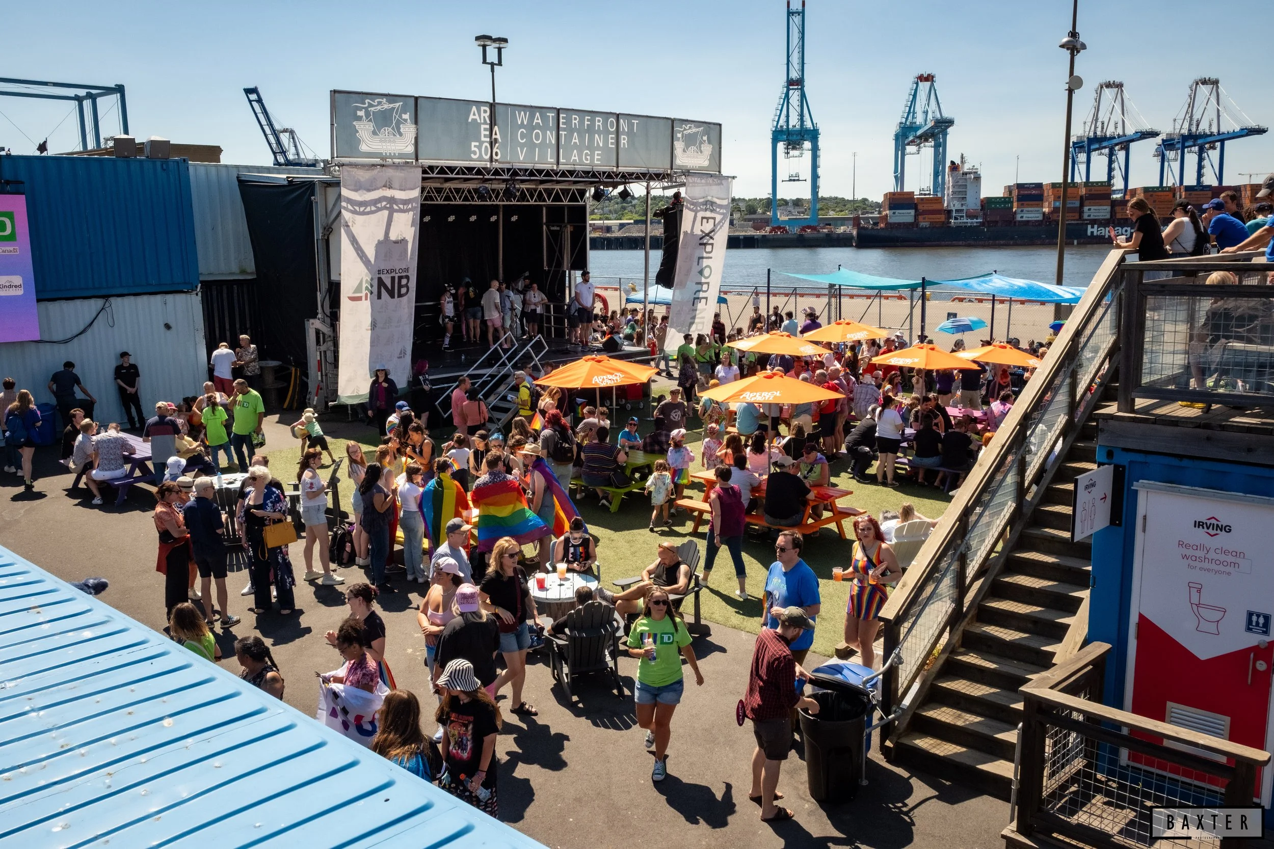Crowd gathered at outdoor event near waterfront with stage, orange umbrellas, and shipping containers in the background.