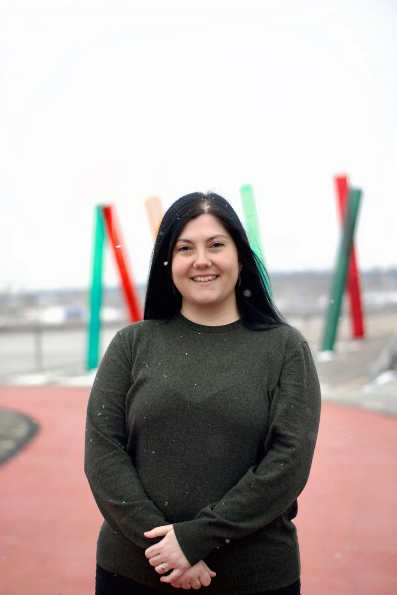 A person with long black hair smiling at the camera outdoors with colorful modern art sculptures in the background.