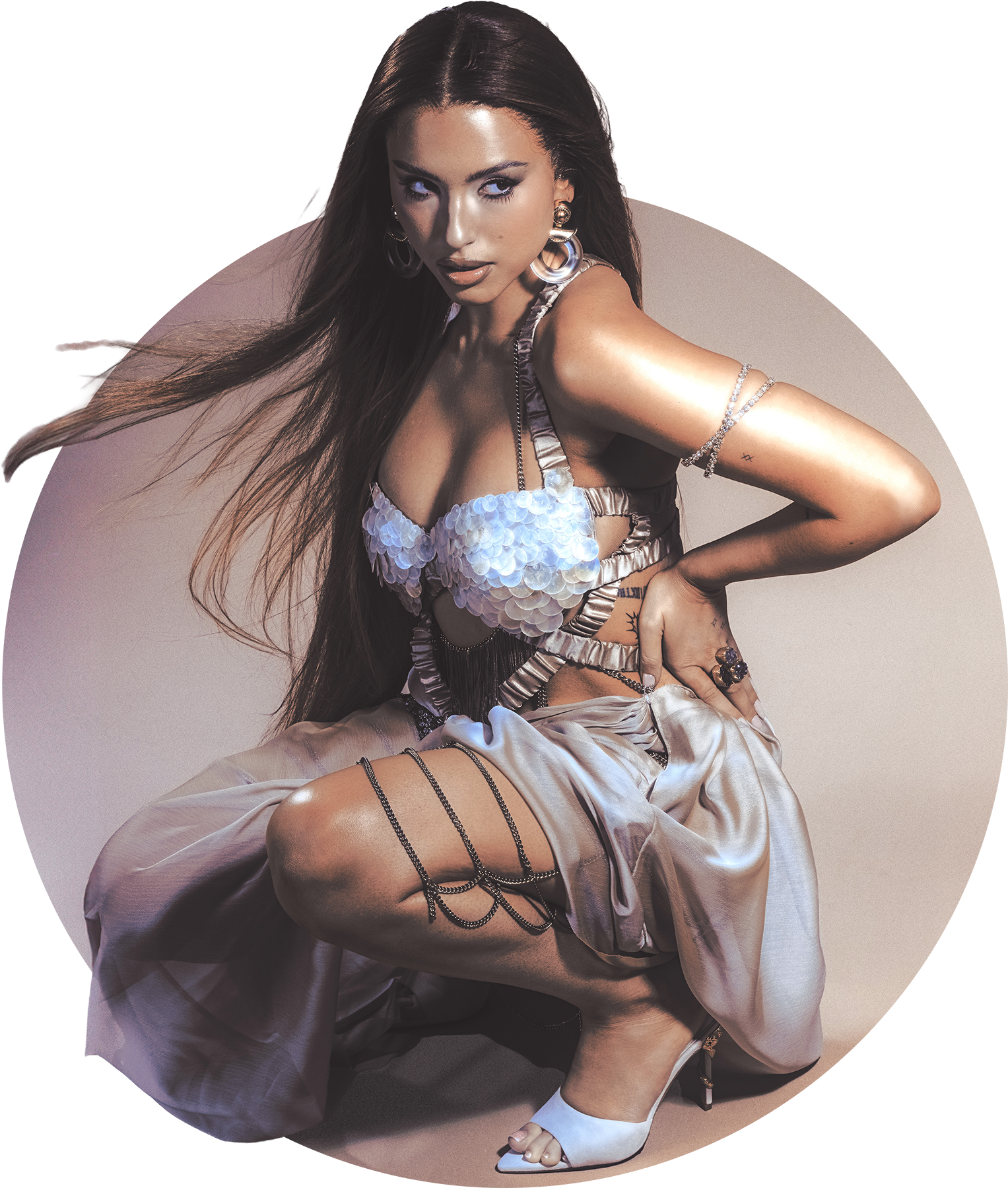 A woman with long brown hair posed sitting on the floor, wearing a shiny, feathered top, satin wide-leg pants, white high heels, and jewelry, against a plain background.