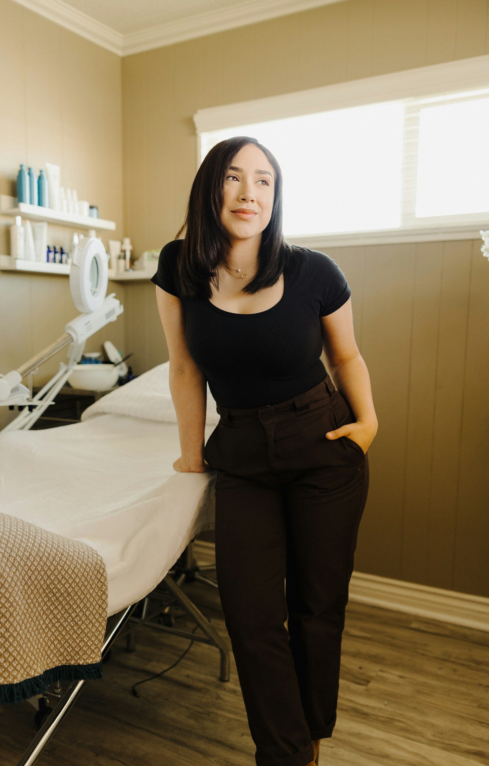 A woman with shoulder-length dark hair wearing a black t-shirt and black pants standing next to a bed in a room with beige walls and a large window. There are shelves with bottles and skincare products in the background.