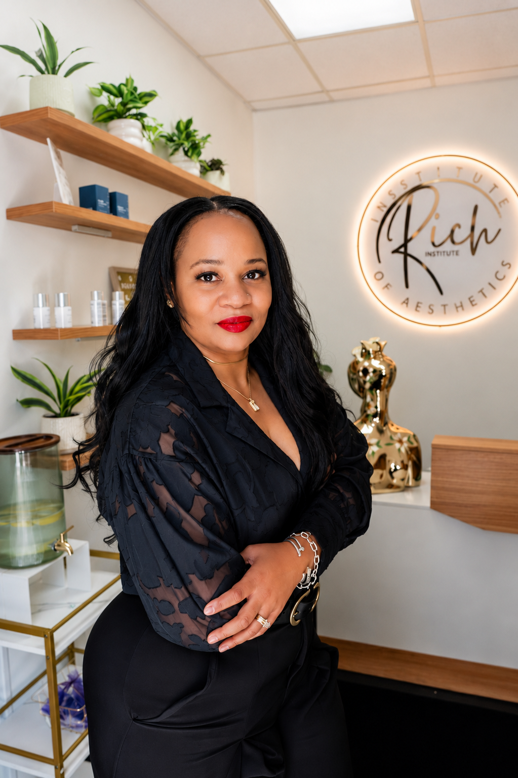A woman with long black hair, wearing a black dress with sheer patterned sleeves, standing inside a modern aesthetic clinic. The clinic has a circular neon sign on the wall that reads 'Rich Institute of Aesthetics,' with shelves of plants and skincare products behind her. She has red lipstick and is wearing jewelry, including a necklace, bracelet, and rings.