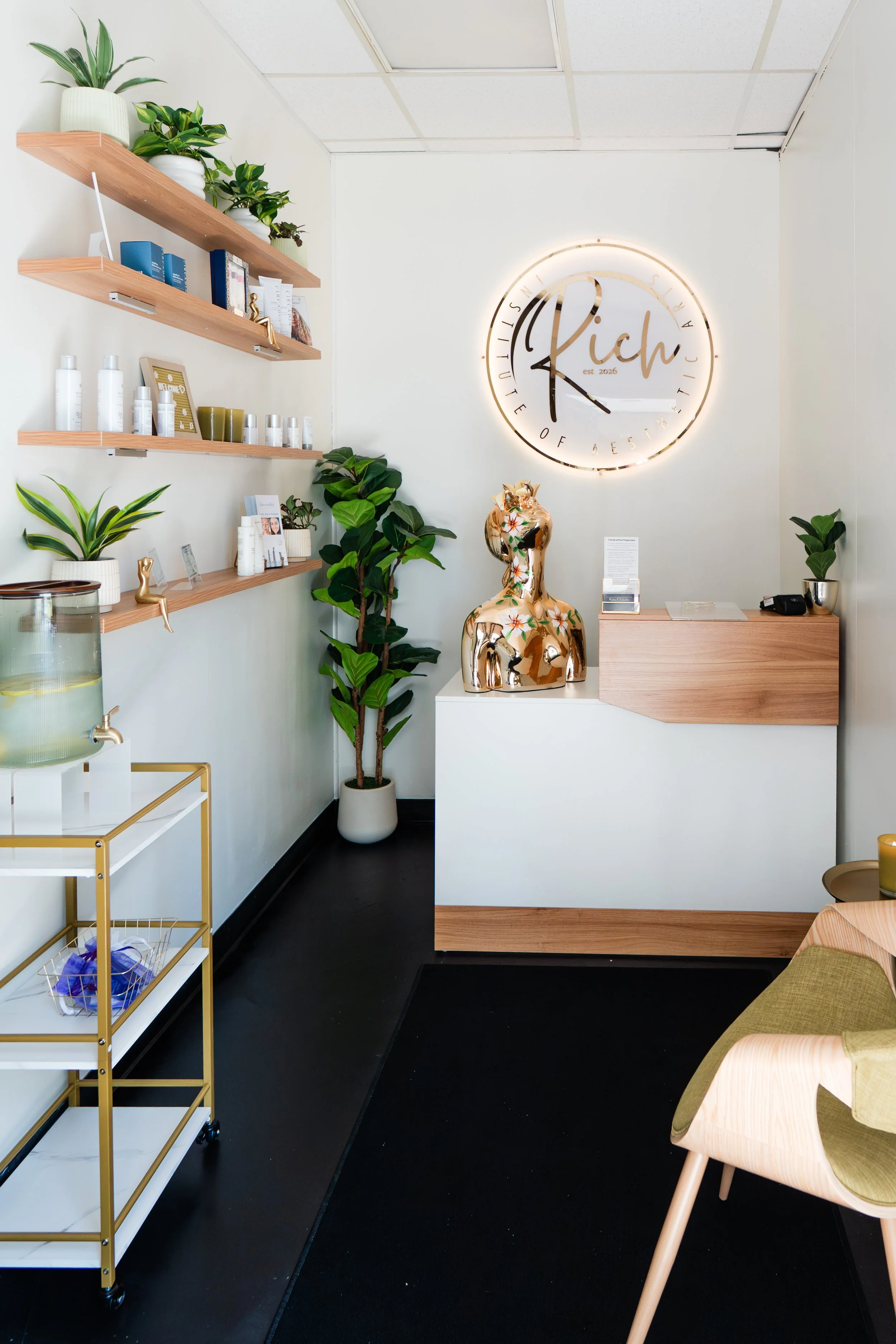 Interior of a modern aesthetic beauty or skincare clinic with white walls, wooden shelves displaying skincare products, potted plants, and a large clock with a neon sign reading 'Rich' on the wall. There is a decorative gold sculpture of a female torso on a white stand and a reception area with a desk.