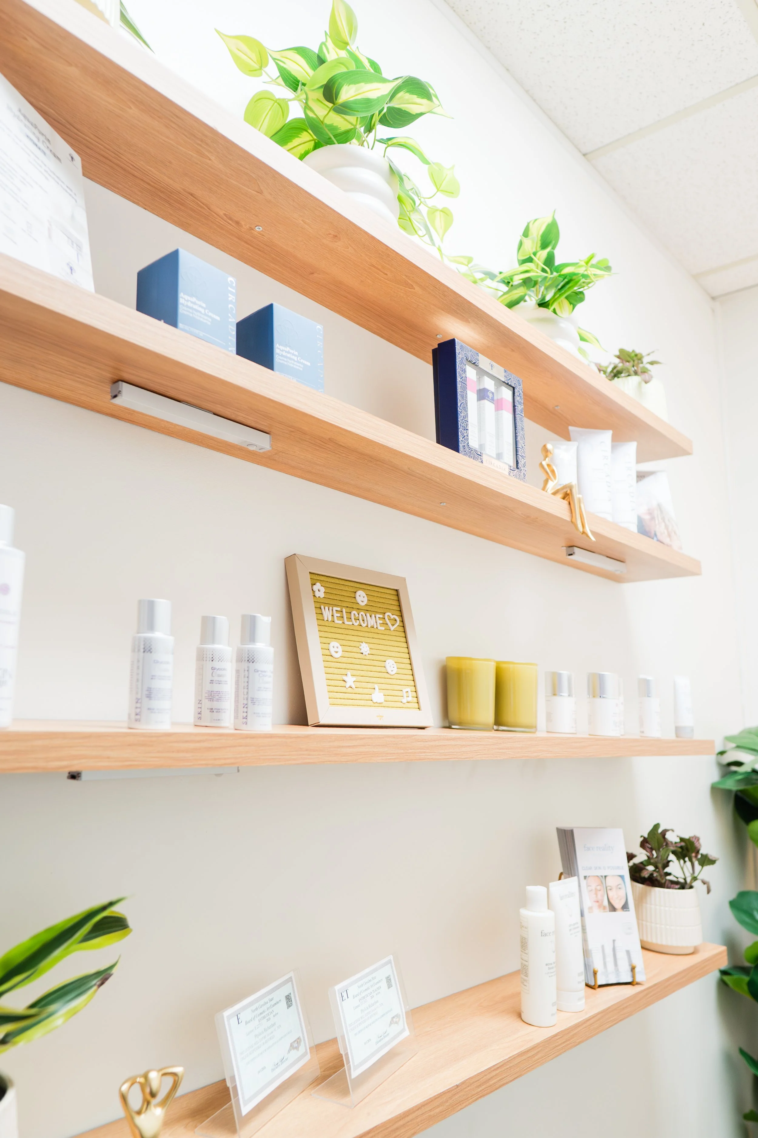 Wooden shelves decorated with green potted plants and skincare products, framed pictures, and a greeting sign.