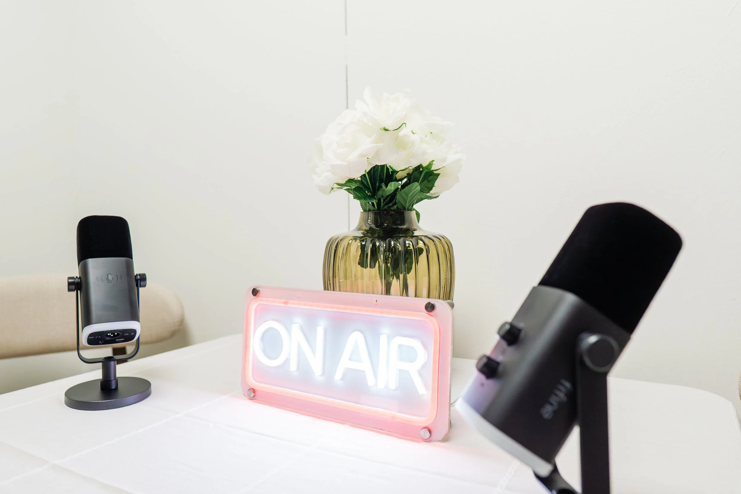 A table with a potted white flower in a gold vase, two black microphones, and a pink neon "ON AIR" sign.